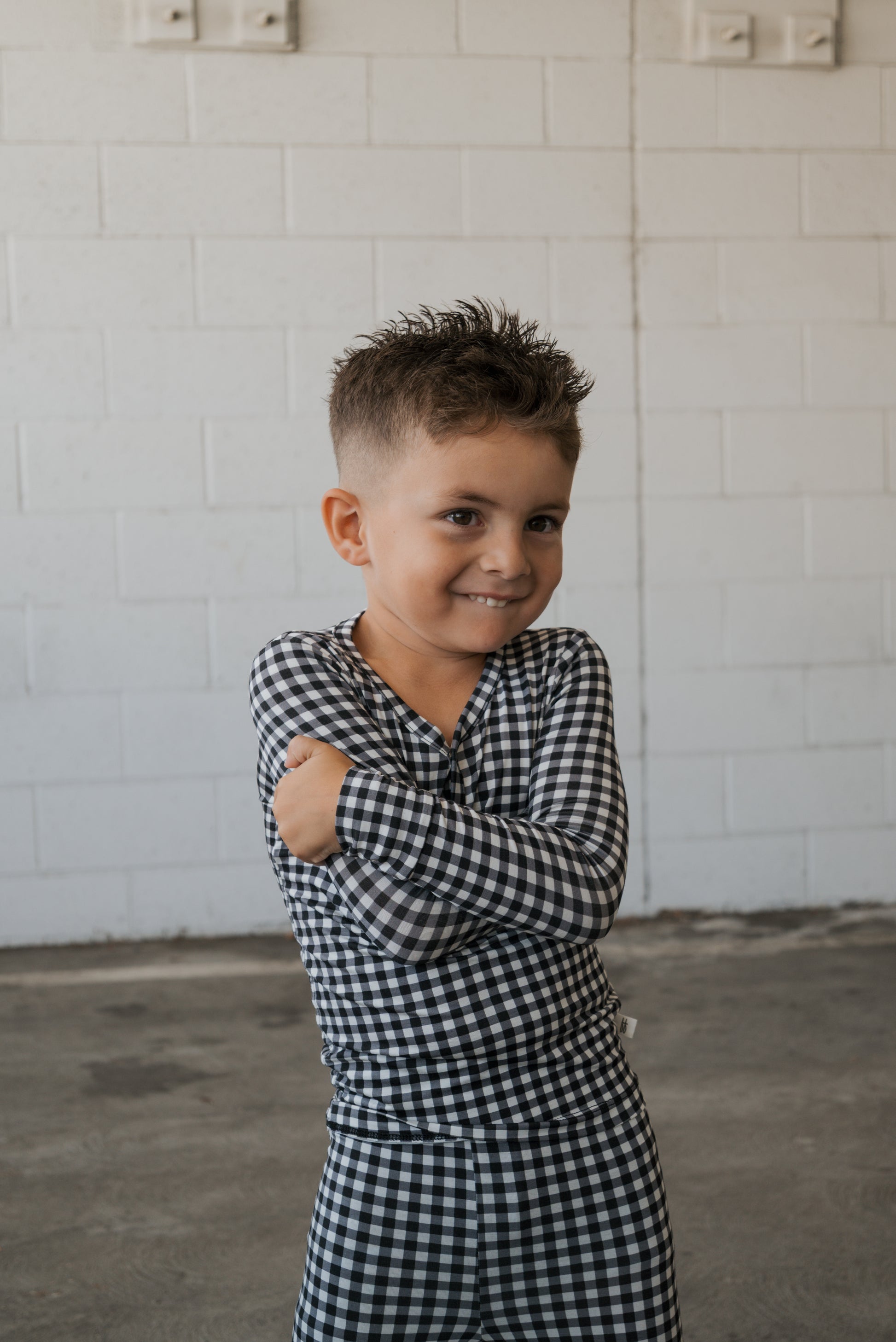 A young boy smiles with his arms crossed, wearing the forever french baby Bamboo Two Piece Pajamas in Black Gingham, standing in front of a white brick wall.