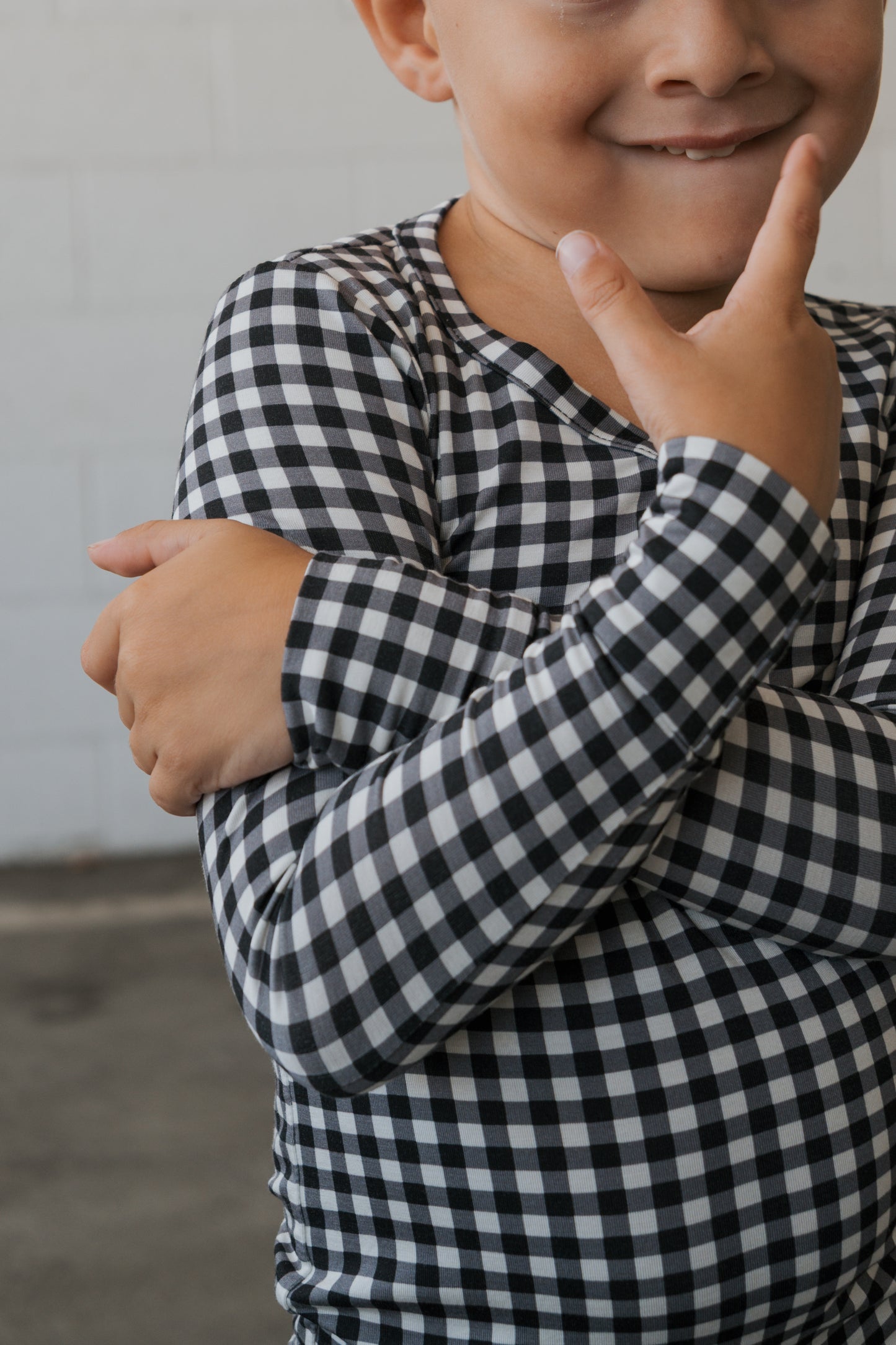 Dressed in the forever french baby Bamboo Two Piece Pajamas in classic Black Gingham, a child smiles brightly—posing playfully with one hand on their chin and the other arm crossed over their chest—to highlight the cozy comfort and effortless style of these breathable pajamas, perfect for everyday adventures.