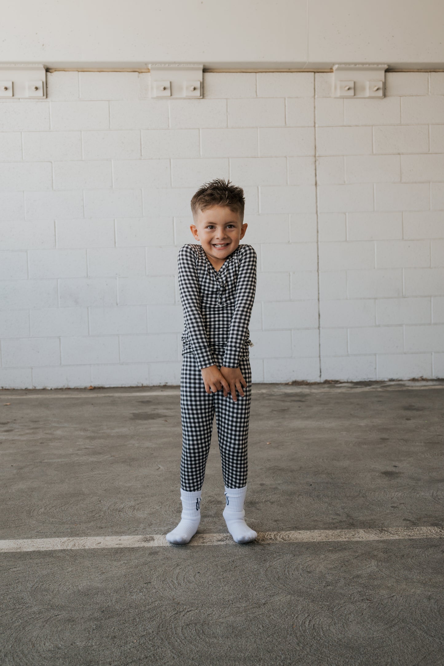 A young child wearing forever french baby’s Bamboo Two Piece Pajamas in Black Gingham and white socks smiles while standing in an empty indoor space with a white brick wall background.