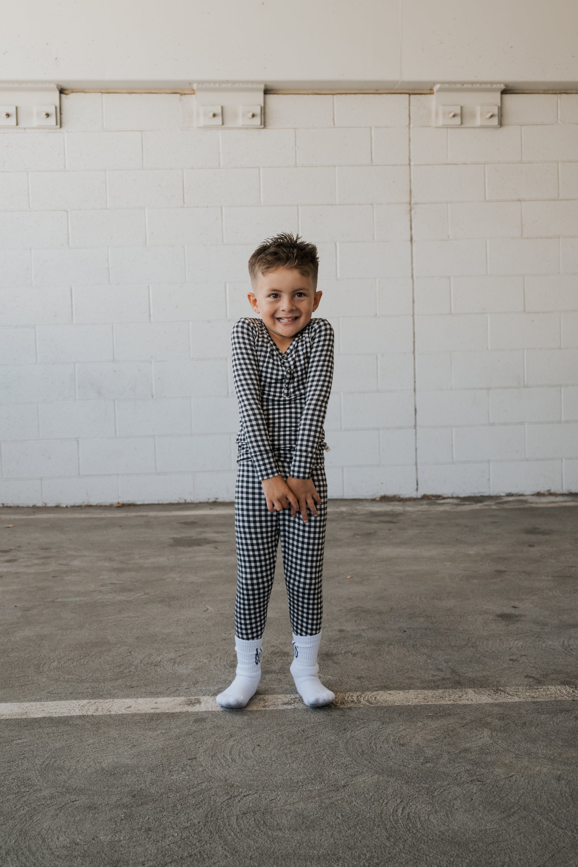 A young child wearing forever french baby’s Bamboo Two Piece Pajamas in Black Gingham and white socks smiles while standing in an empty indoor space with a white brick wall background.