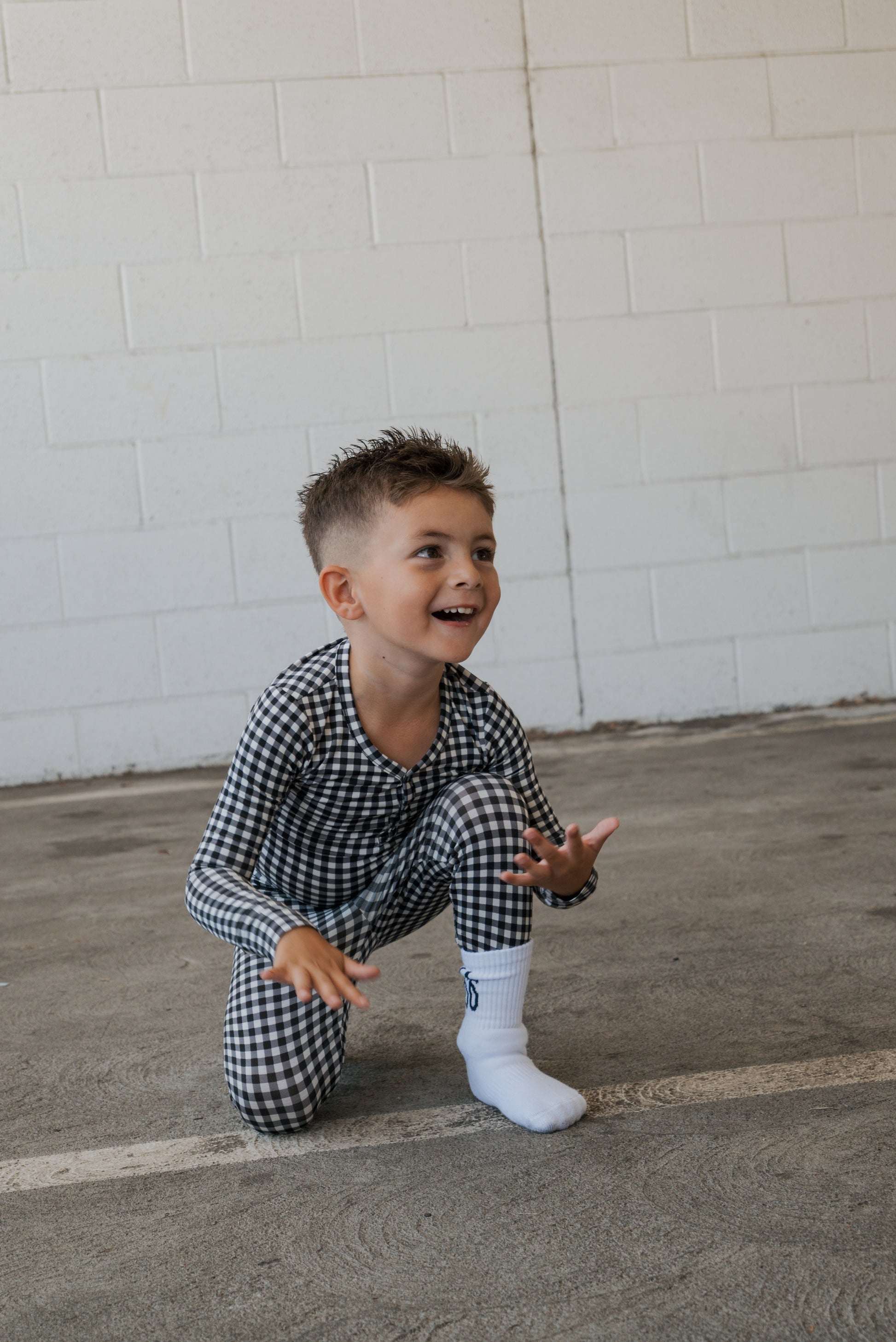 A young boy wearing the forever french baby Bamboo Two Piece Pajamas in Black Gingham crouches and smiles on a concrete floor near a white brick wall.