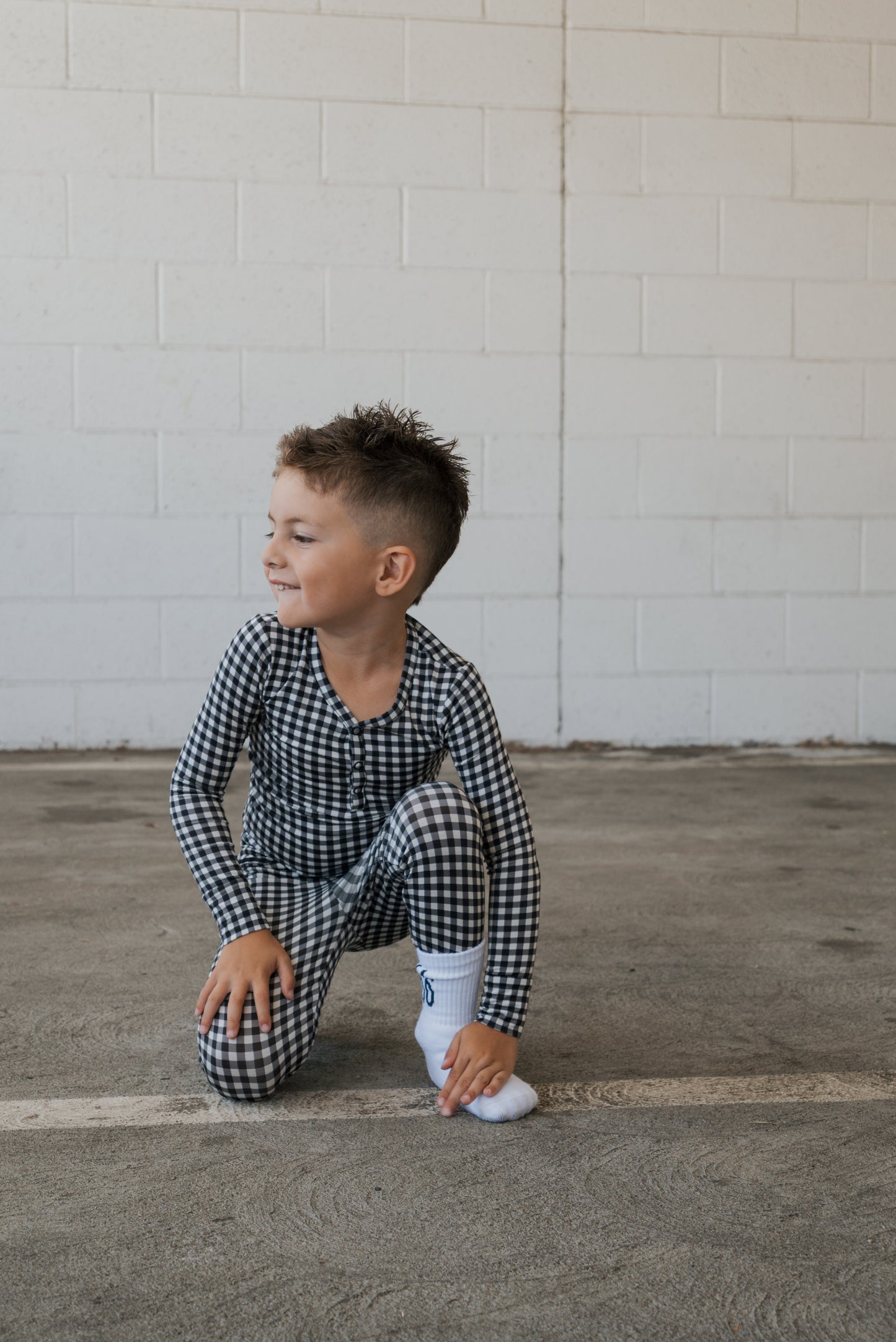 A young child wearing the Bamboo Two Piece Pajamas in Black Gingham from forever french baby crouches on a concrete floor, smiling and looking to the side, with a white brick wall in the background.