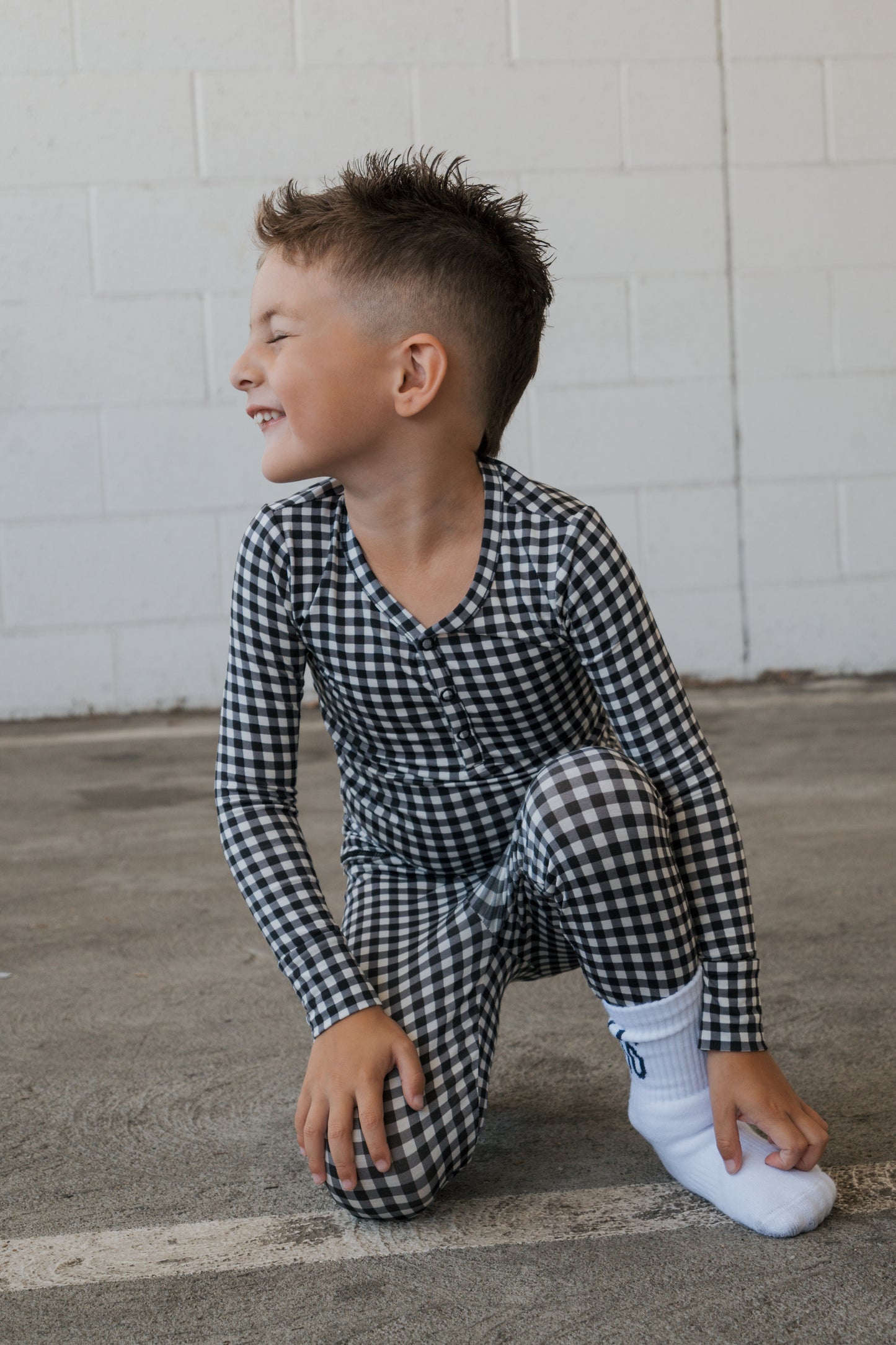 A young boy wearing forever french baby’s Bamboo Two Piece Pajamas in Black Gingham with white socks kneels on the ground, smiling and looking to the side against a light brick wall background.