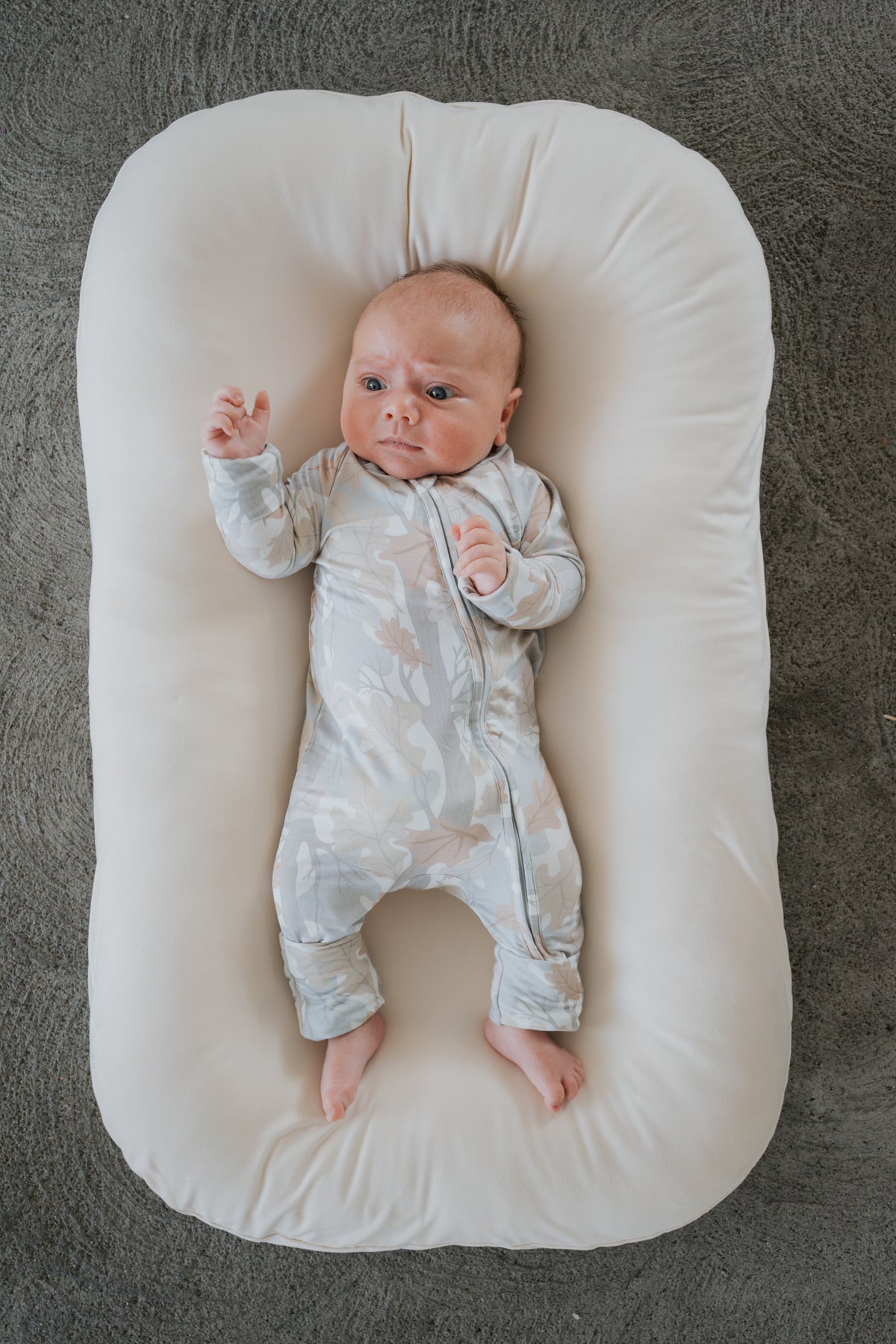 Dressed in forever french baby Bamboo Zip Pajamas in the Woodland print, a baby lies on a soft, white cushion with one arm raised, gazing upward against a textured gray backdrop.