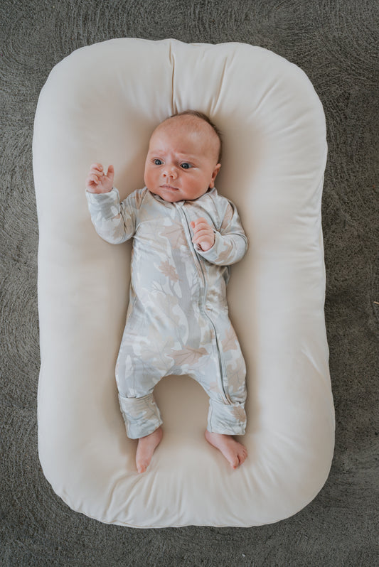 Dressed in forever french baby Bamboo Zip Pajamas in the Woodland print, a baby lies on a soft, white cushion with one arm raised, gazing upward against a textured gray backdrop.