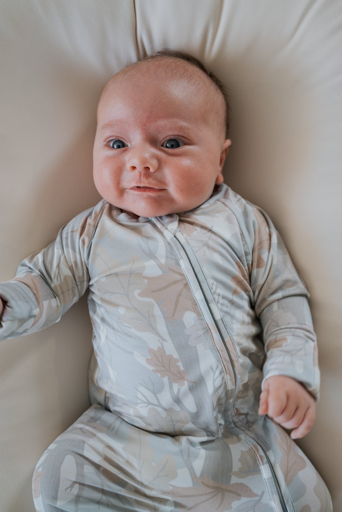 A baby in the forever french baby Bamboo Zip Pajamas | Woodland, a patterned, hypoallergenic zip-up onesie, lies on a light-colored cushion, looking up with wide eyes and a slight smile.