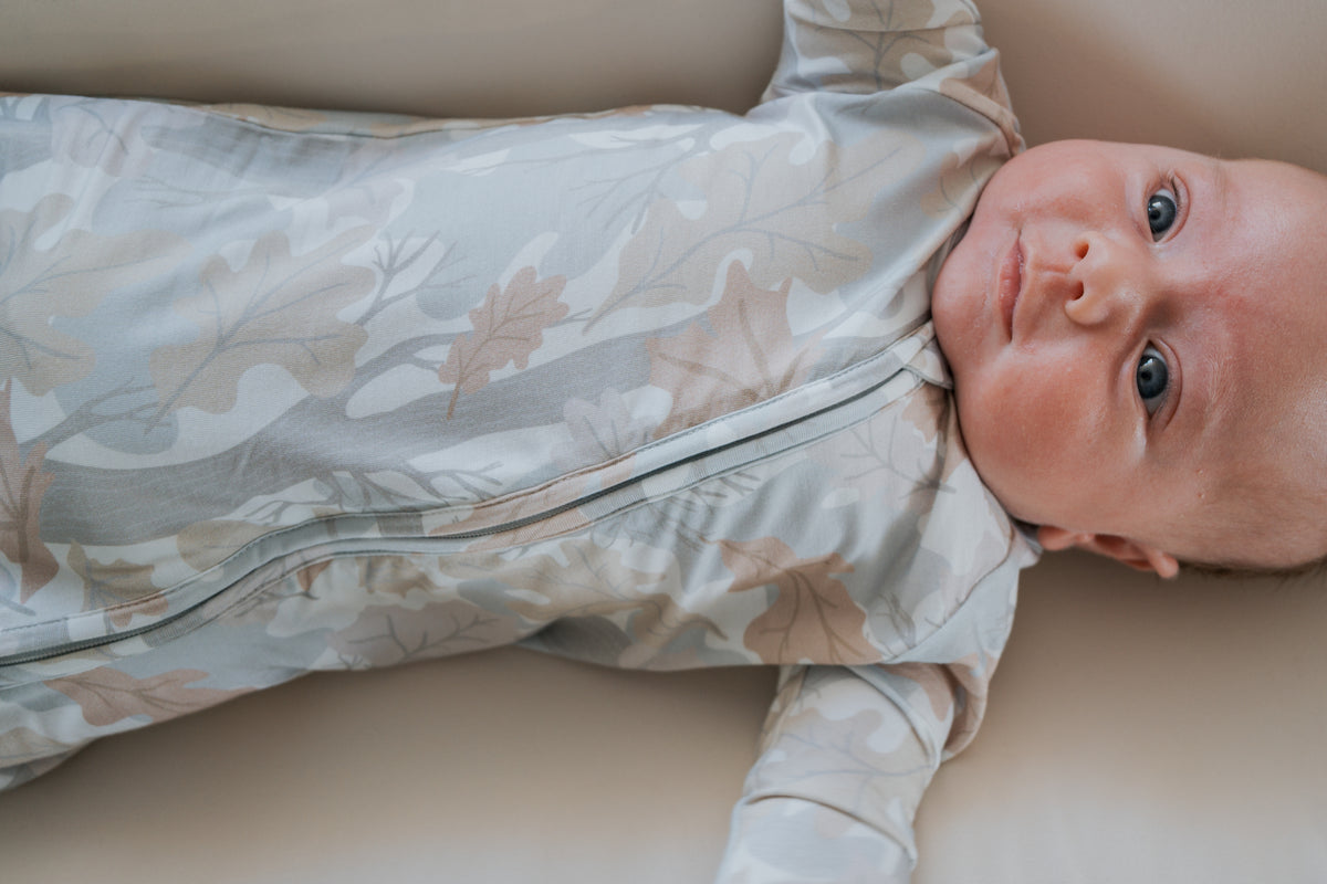 A baby is lying on a light surface, dressed in forever french baby’s Bamboo Zip Pajamas | Woodland—hypoallergenic sleepwear featuring a nature-inspired leaf pattern. The baby gazes upward with wide blue eyes and a calm expression.