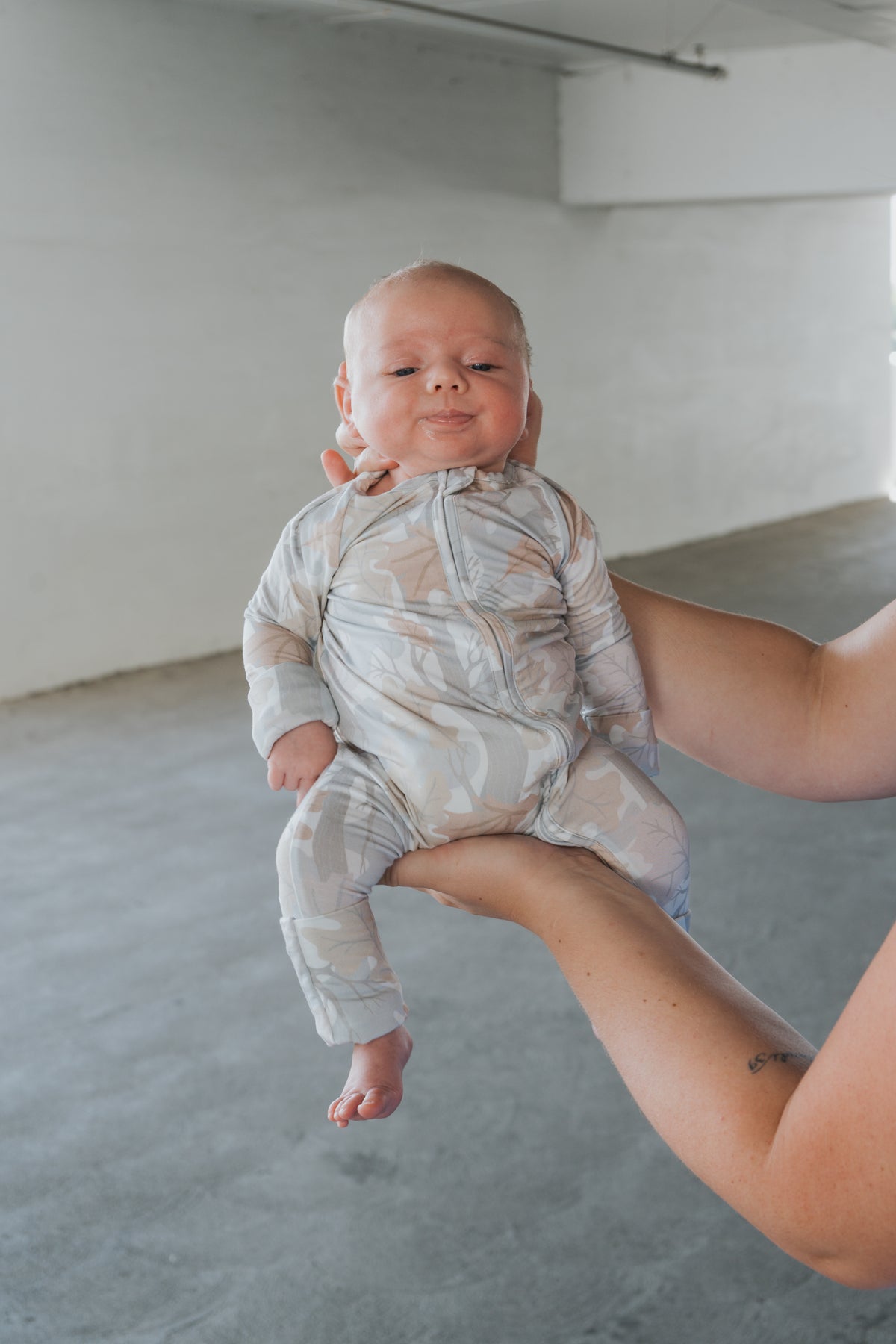 An adult holds up a baby dressed in forever french baby’s Bamboo Zip Pajamas | Woodland—a beige, patterned onesie—inside a bright, empty room with gray floors and white walls.