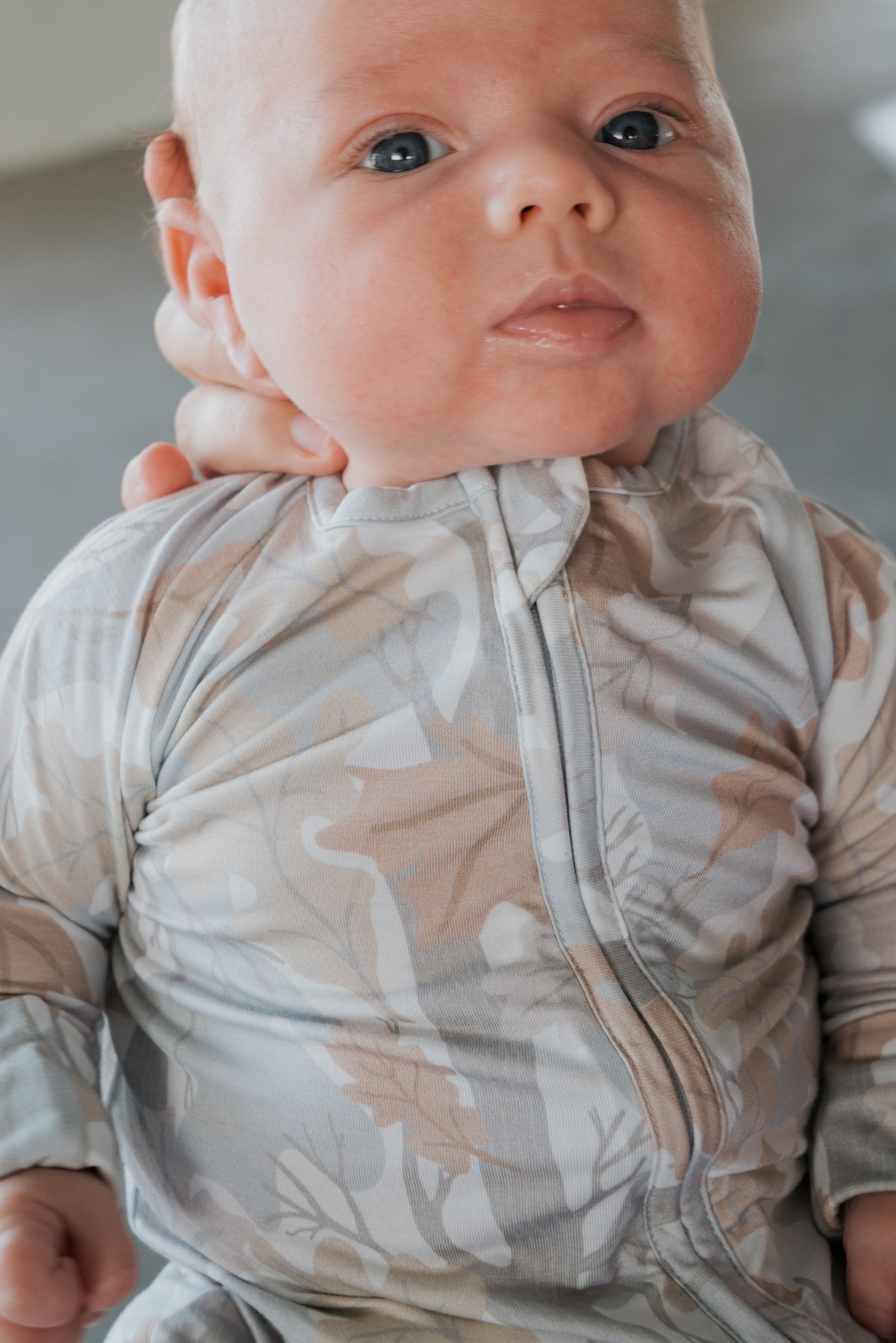 A close-up of a baby wearing forever french baby’s Bamboo Zip Pajamas in the Woodland pattern, featuring a delicate nature-inspired design. An adult’s hand gently supports the baby’s head and neck.