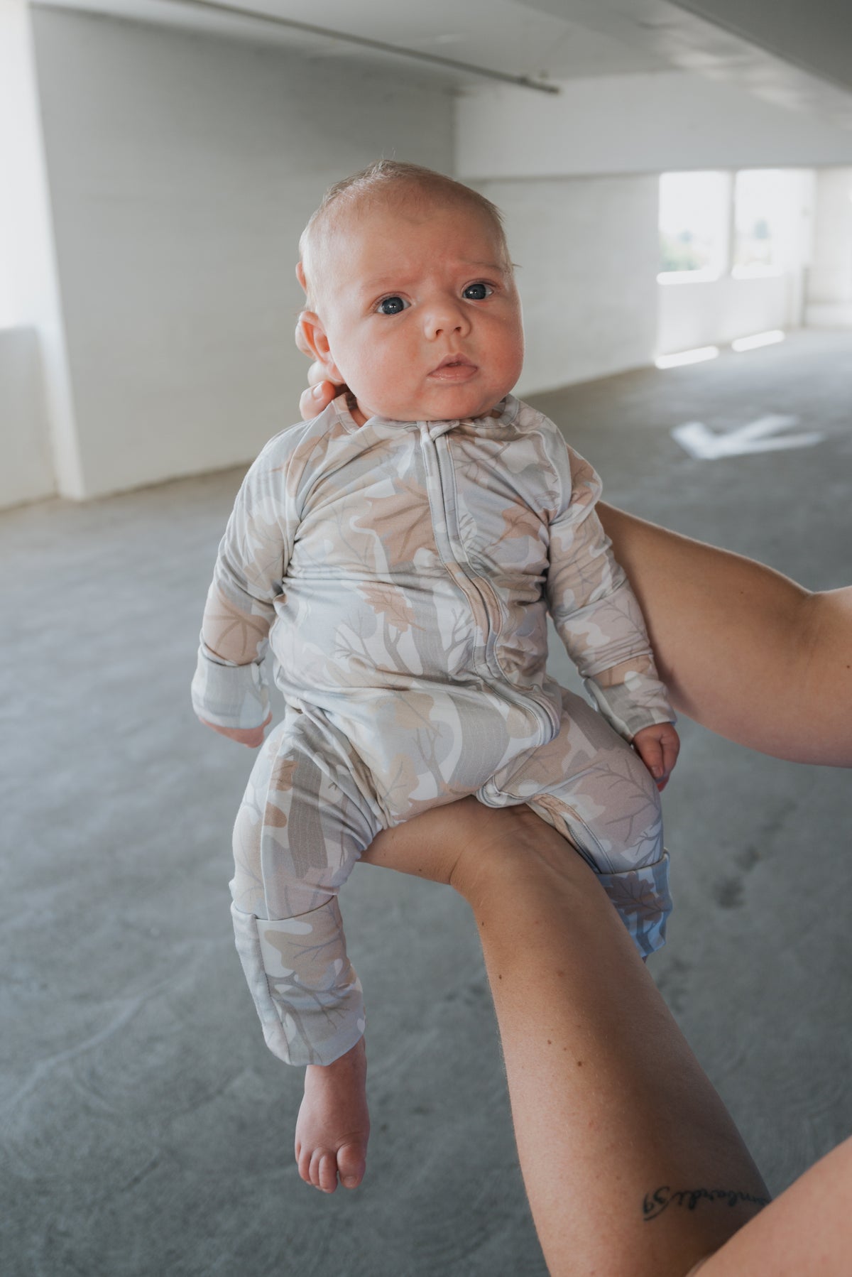 A person holds a baby dressed in forever french baby’s Bamboo Zip Pajamas | Woodland, a light-colored, patterned onesie made from hypoallergenic fabric. The baby looks slightly surprised. The background is an empty, bright indoor space with white walls and windows.