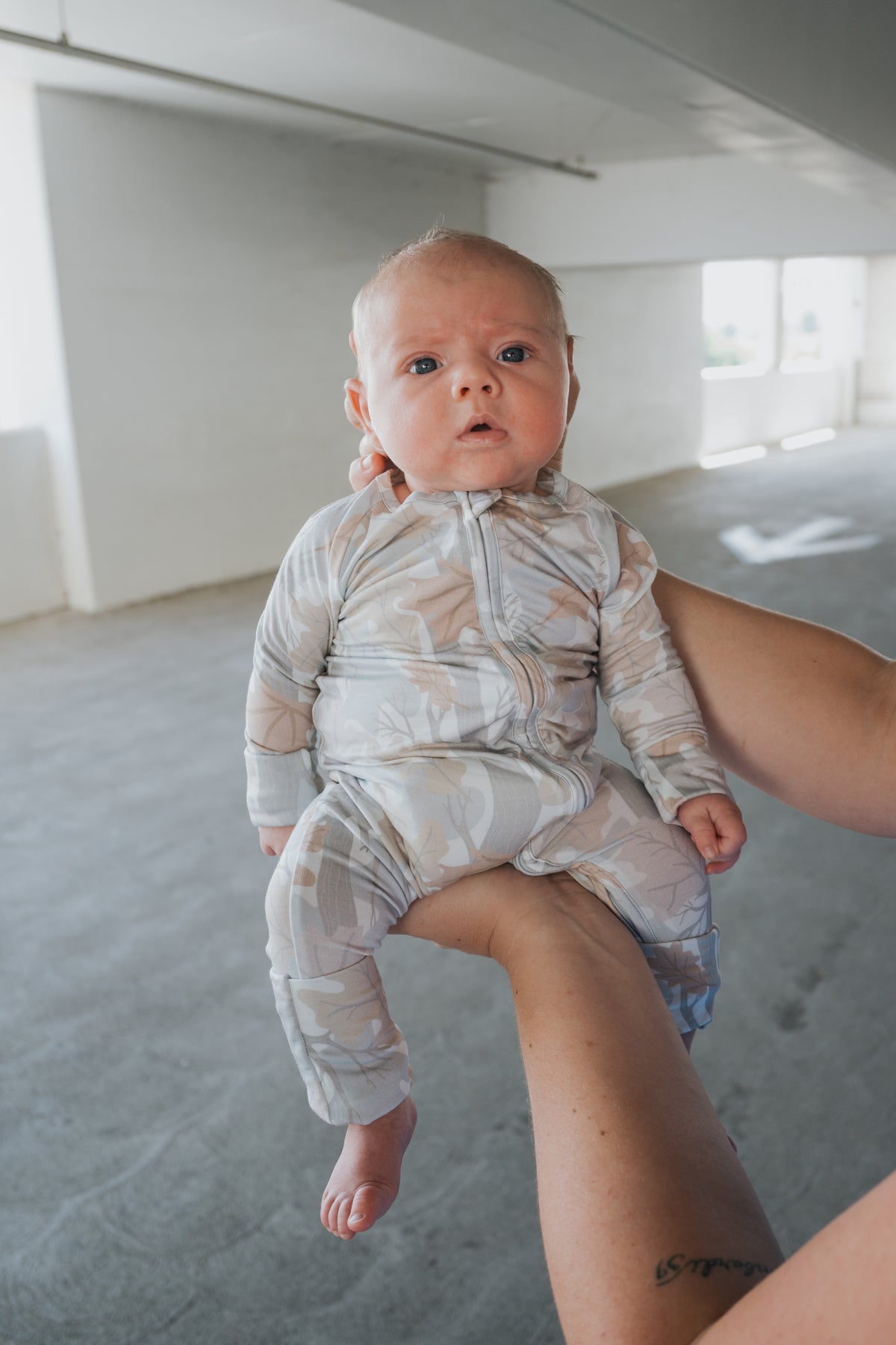 A baby wearing forever french baby’s Bamboo Zip Pajamas in the Woodland print—a soft, hypoallergenic onesie—is held upright by an adult’s arm in a bright, empty indoor space. The baby gazes curiously at the camera.
