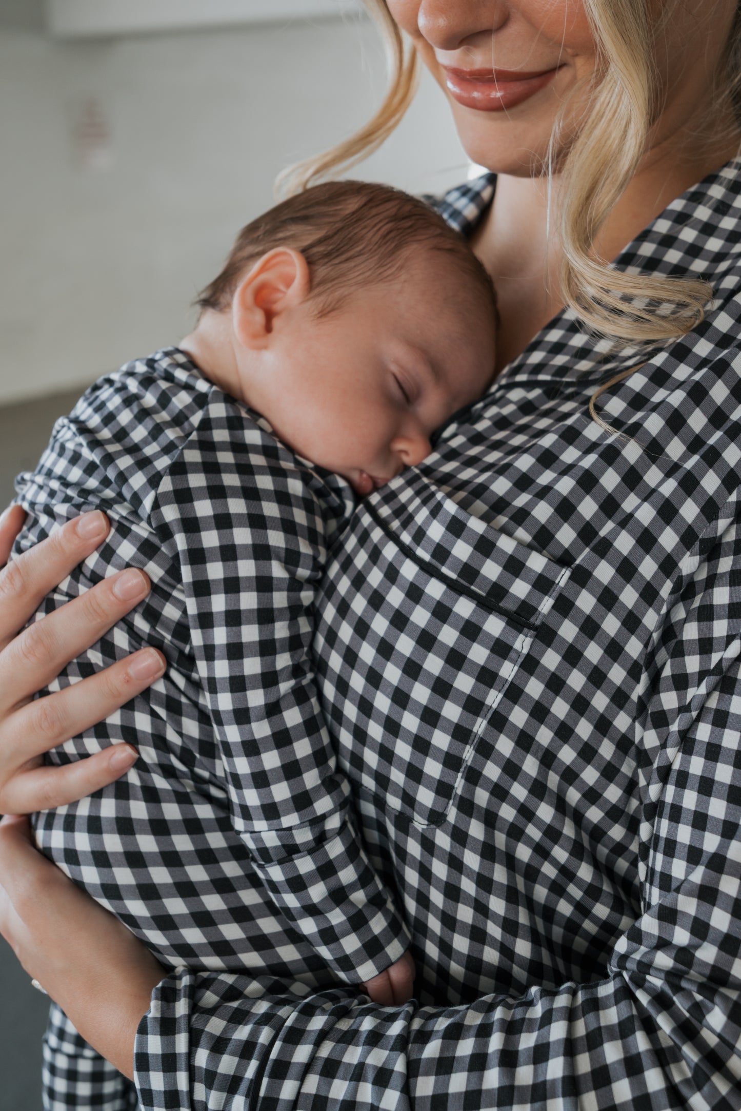 A woman smiles softly while holding a sleeping baby on her chest. Both are wearing matching black gingham outfits, with the woman dressed in cozy wide leg lounge pants from the forever french baby Women’s Bamboo Button Up Lounge Set in Black Gingham. Their faces are close together as the baby rests peacefully.