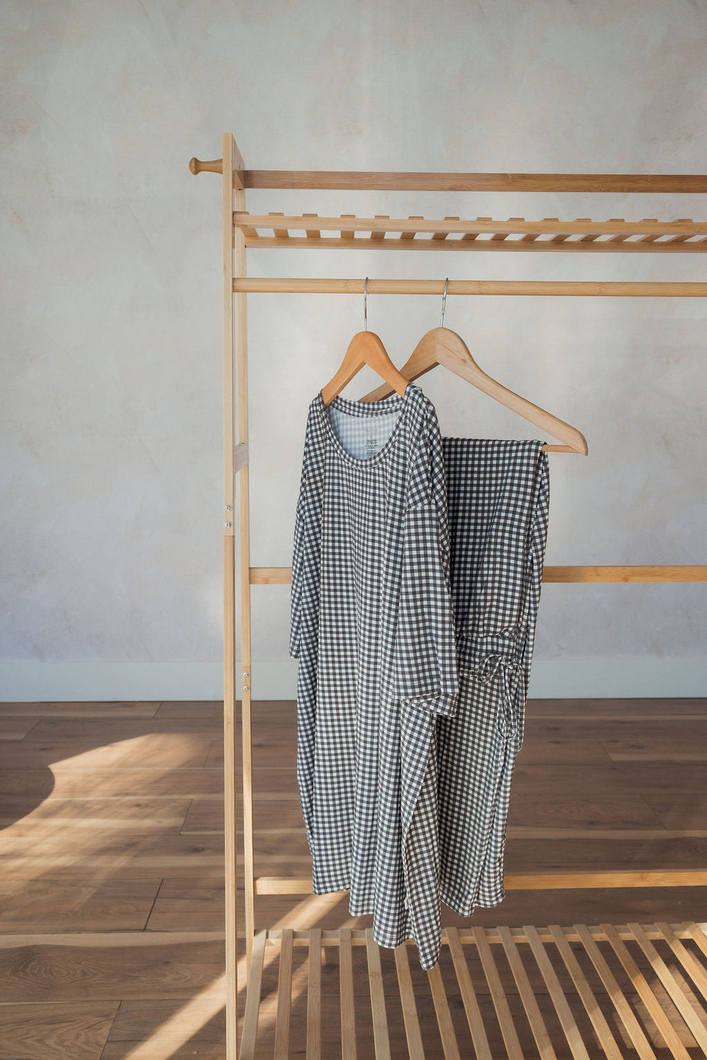 A wooden rack displays two wooden hangers, one with forever french baby Men's Short Sleeve Pajamas in Green Gingham—perfect as comfy, hypoallergenic sleepwear. The rack sits on a wood floor against a light wall.