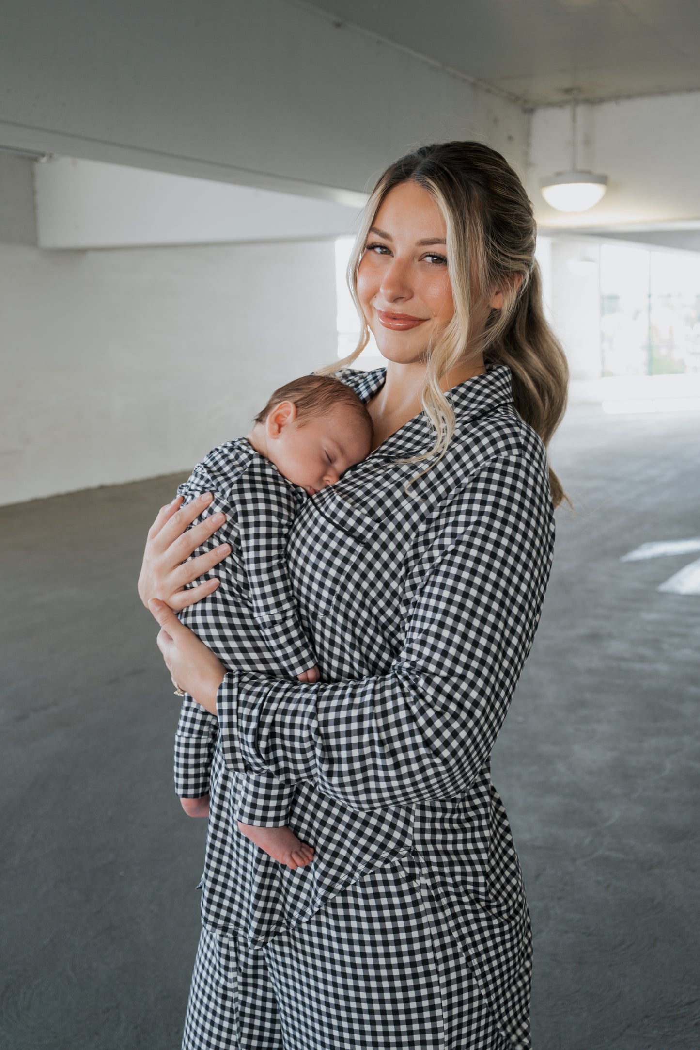 A woman wearing the forever french baby Women’s Bamboo Button Up Lounge Set in Black Gingham smiles softly while holding a sleeping baby dressed in matching black gingham bamboo clothing, both in a bright indoor space.