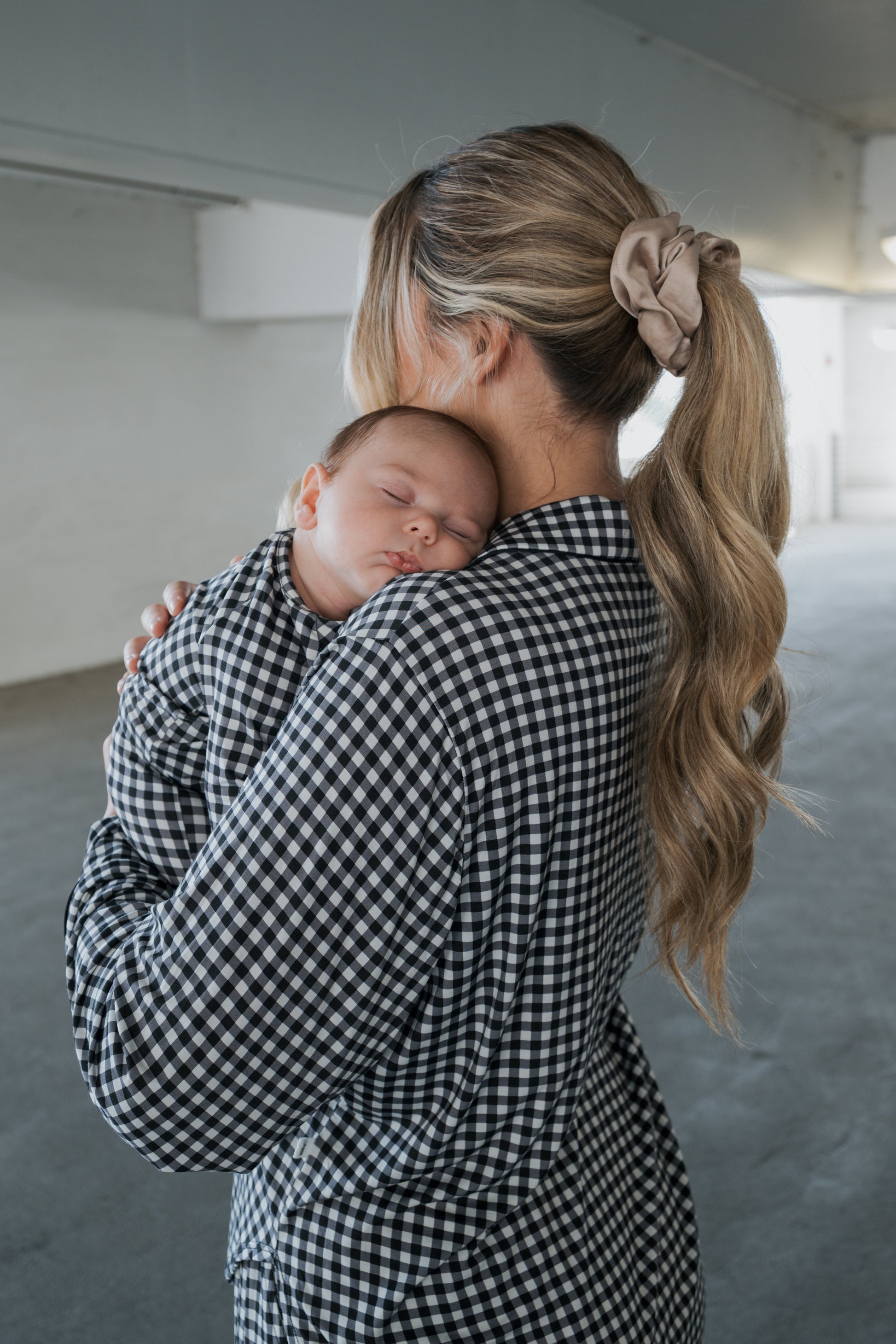 A woman with long blond hair in a ponytail holds a sleeping baby on her shoulder. Both are dressed in matching black gingham bamboo clothing from forever french baby, with the woman wearing the Women’s Bamboo Button Up Lounge Set. They are indoors, softly lit from the right, creating a cozy and stylish look.