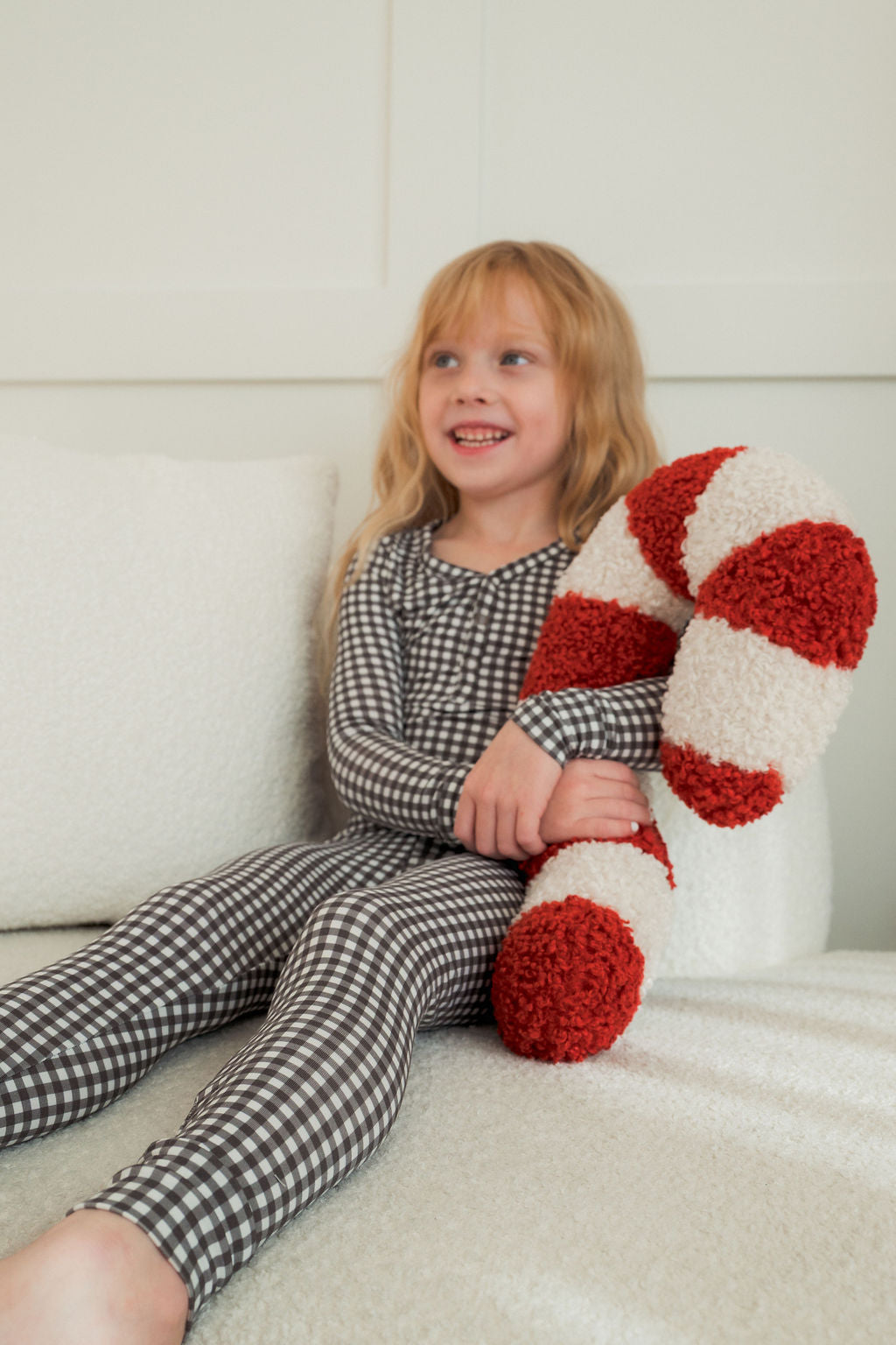 A young child wearing forever french baby’s Bamboo Two Piece Pajamas in Green Gingham sits on a cream couch, smiling while holding a large red-and-white striped plush candy cane.