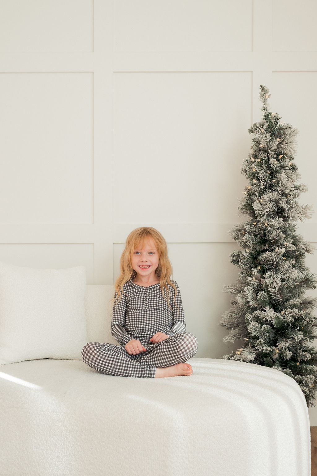 A young girl with long blonde hair smiles, sitting cross-legged on a white bed in forever french baby’s Bamboo Two Piece Pajamas in Green Gingham. Beside her, a small snowy artificial Christmas tree stands against a light wall.