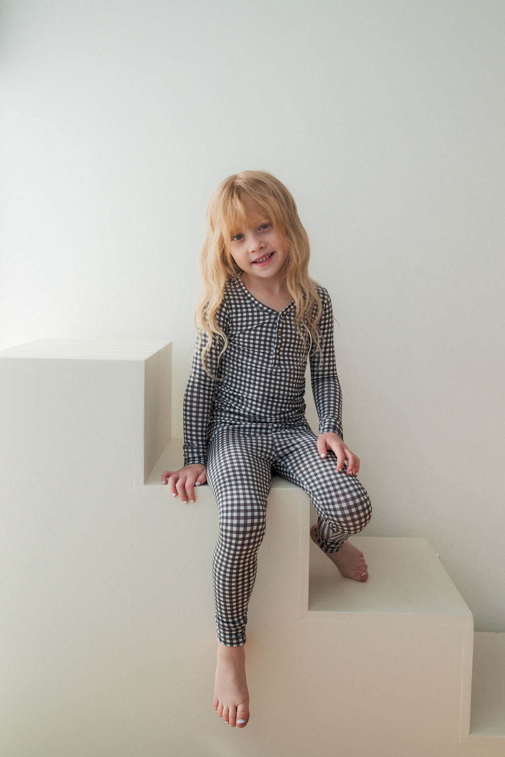 A young child with long blonde hair, barefoot and smiling at the camera, wears forever french baby’s Bamboo Two Piece Pajamas in Green Gingham while sitting on white block steps against a plain white background.