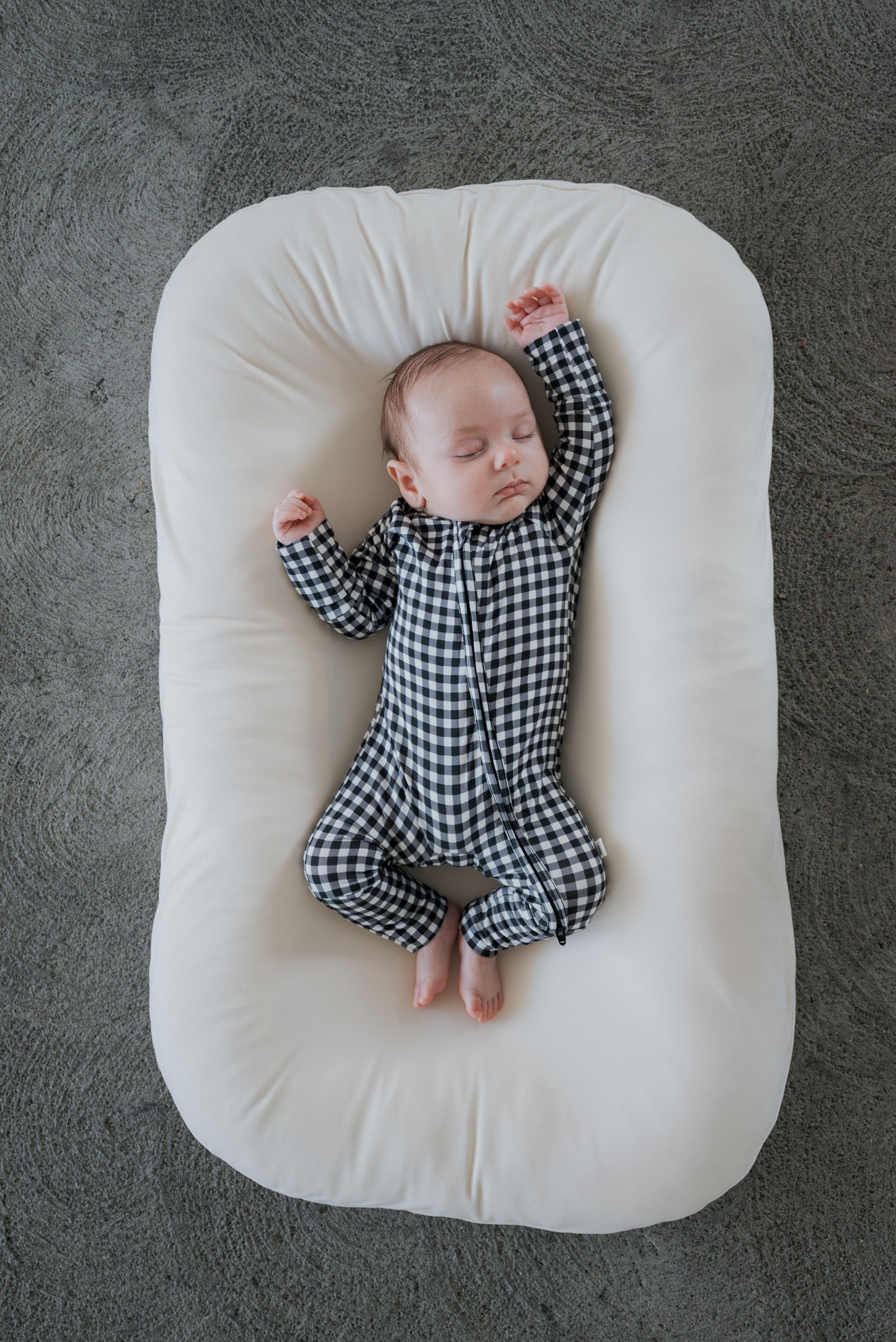 A baby wearing forever french baby’s Bamboo Zip Pajamas in Black Gingham sleeps peacefully on a white cushioned pad, lying on a gray textured surface.