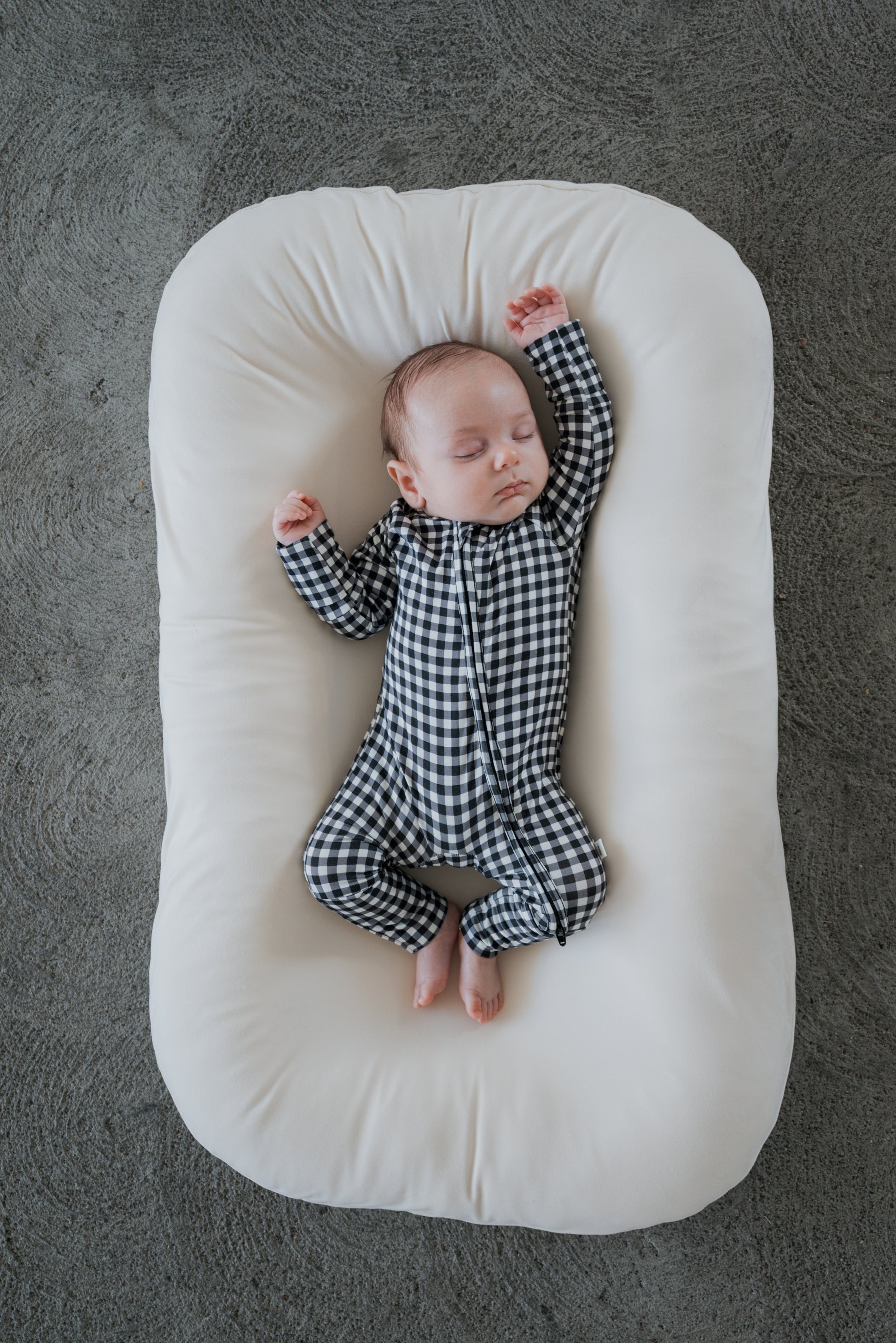 A baby wearing forever french baby’s Bamboo Zip Pajamas in Black Gingham sleeps peacefully on a white cushioned pad, lying on a gray textured surface.