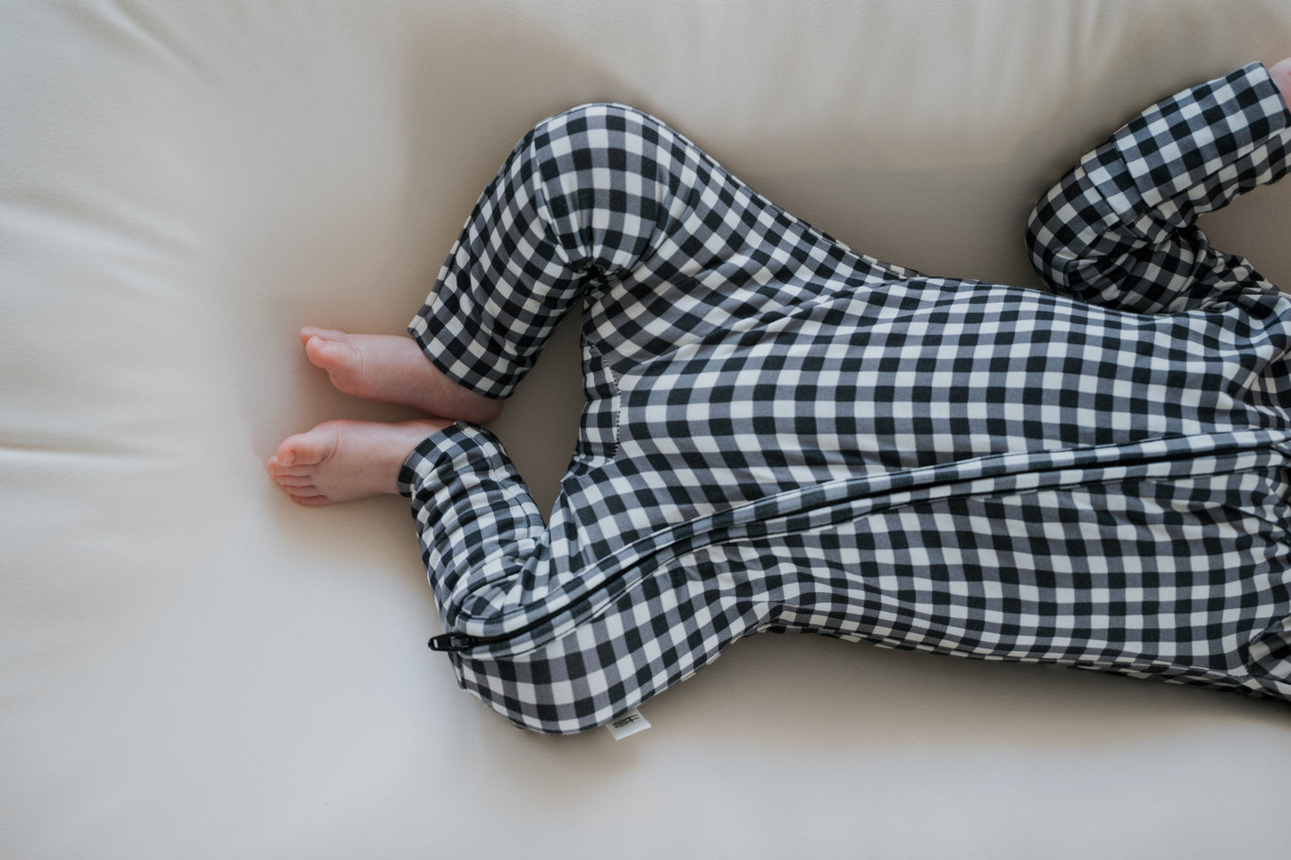A baby wearing forever french baby’s Bamboo Zip Pajamas in Black Gingham, made from breathable fabric, lies on a soft, light-colored surface with only the lower half of their body visible.