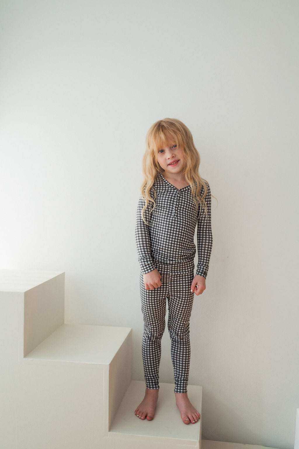 A young child with long blonde hair stands barefoot on white steps against a plain wall, wearing forever french baby’s Bamboo Two Piece Pajamas in Green Gingham—a breathable, matching long-sleeve top and leggings.