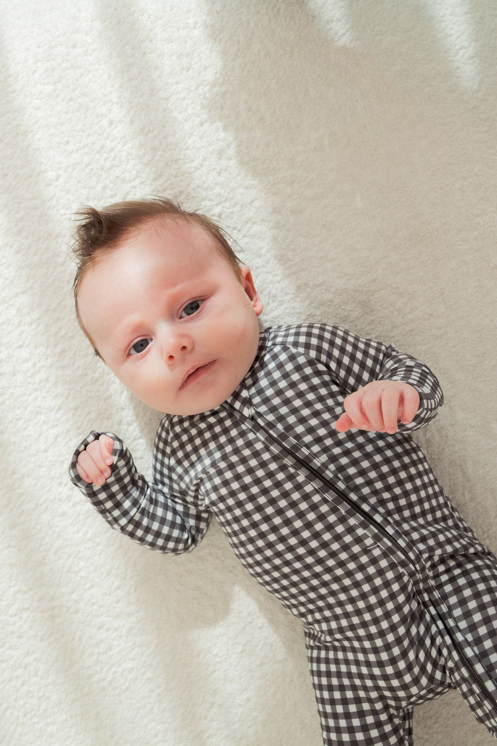 A baby in forever french baby's Bamboo Zip Pajamas in Green Gingham lies on a soft, light-colored blanket, looking up with hands slightly raised.