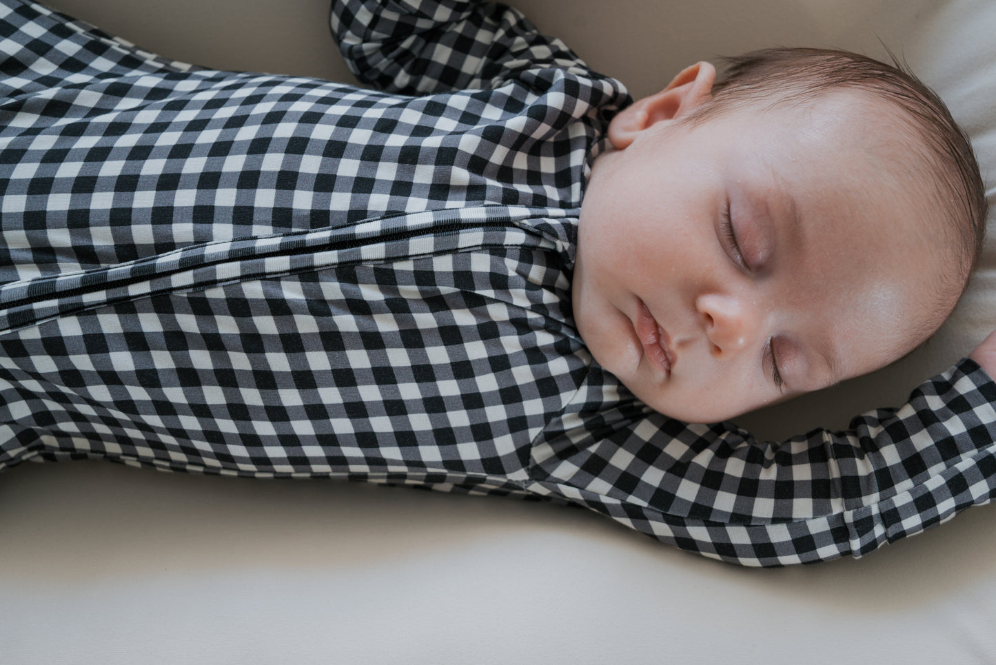 A baby sleeps peacefully on a light surface, wearing forever french baby’s Bamboo Zip Pajamas in Black Gingham, with one arm stretched above their head and eyes closed.