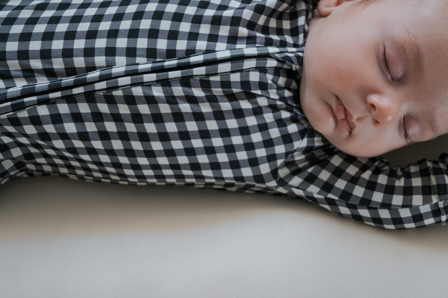 A baby sleeps peacefully, dressed in forever french baby’s Bamboo Zip Pajamas in Black Gingham, lying on a light-colored surface. Only the baby’s face, upper body, and one outstretched arm are visible—highlighting the breathable bamboo sleepwear that keeps them comfortable all night long.