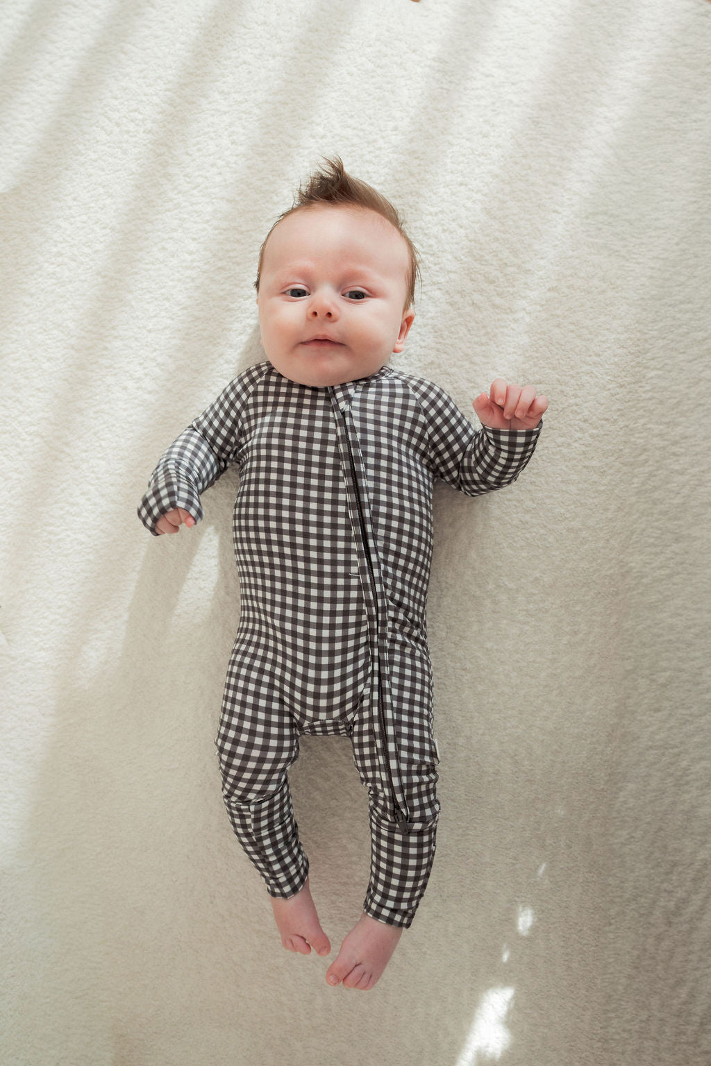 A baby relaxes on a light surface in forever french baby's Bamboo Zip Pajamas in Green Gingham, with gentle sunlight and soft shadows highlighting the breathable, long-sleeved sleepsuit.