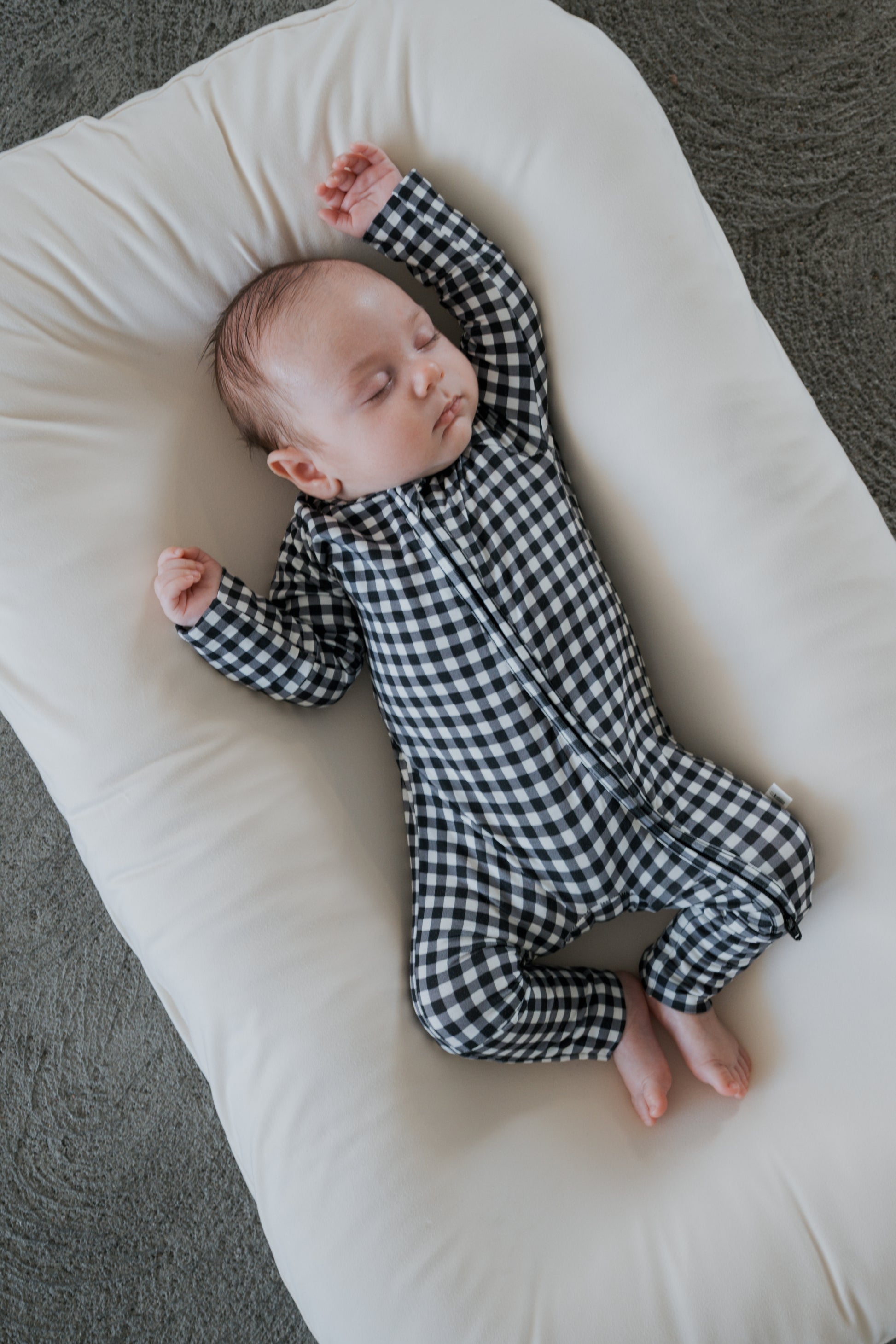 A baby wearing forever french baby Bamboo Zip Pajamas in Black Gingham sleeps on a white cushion with arms raised, lying on a gray textured surface.