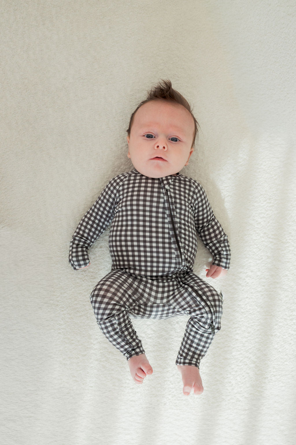 A baby relaxes on a light surface, dressed in forever french baby's Bamboo Zip Pajamas in Green Gingham. The soft, breathable sleepwear keeps them comfy as they gaze calmly upward.