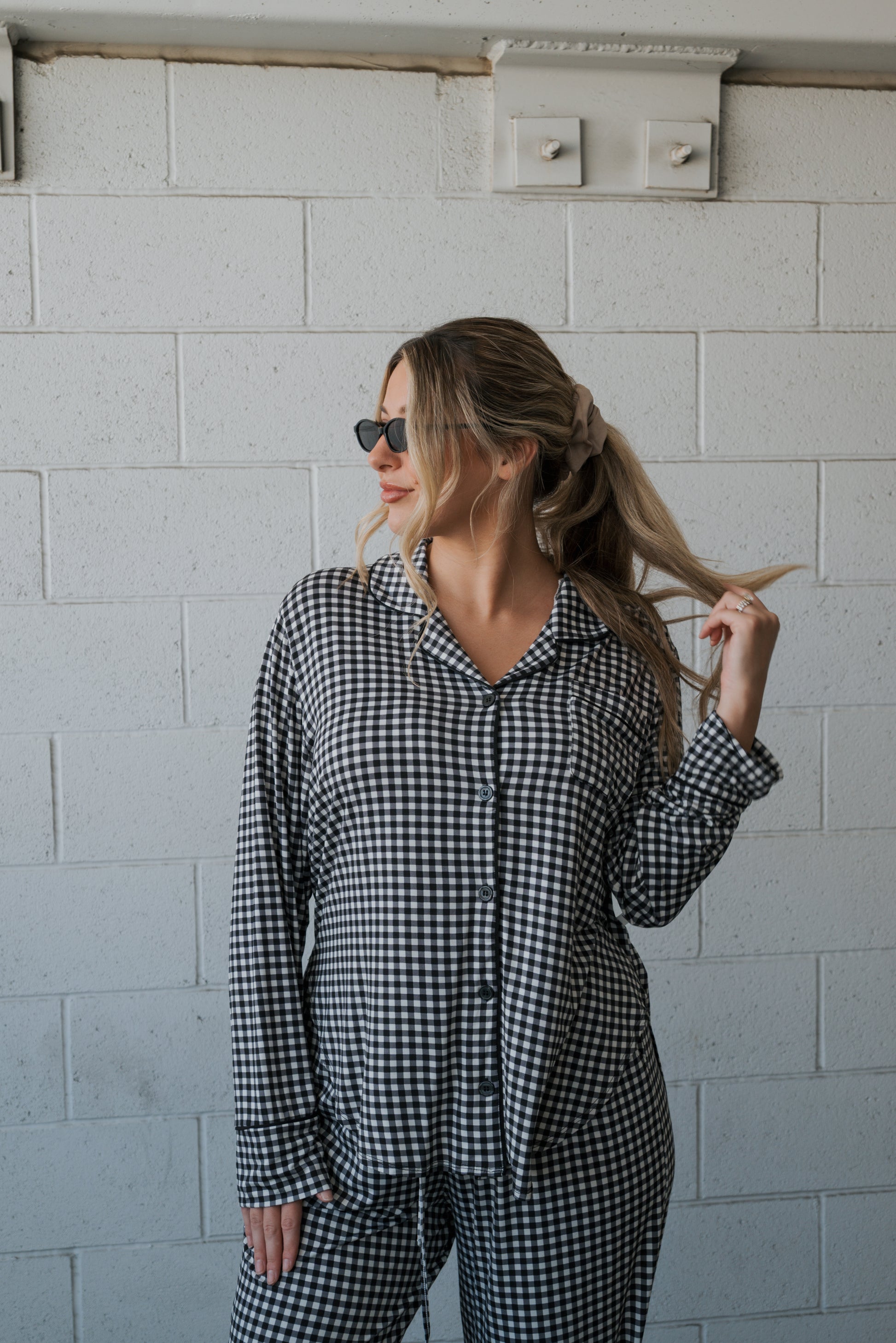 A woman wearing the forever french baby Women’s Bamboo Button Up Lounge Set in Black Gingham with wide leg lounge pants and sunglasses stands against a white brick wall, holding a strand of her long hair with one hand and looking to the side.