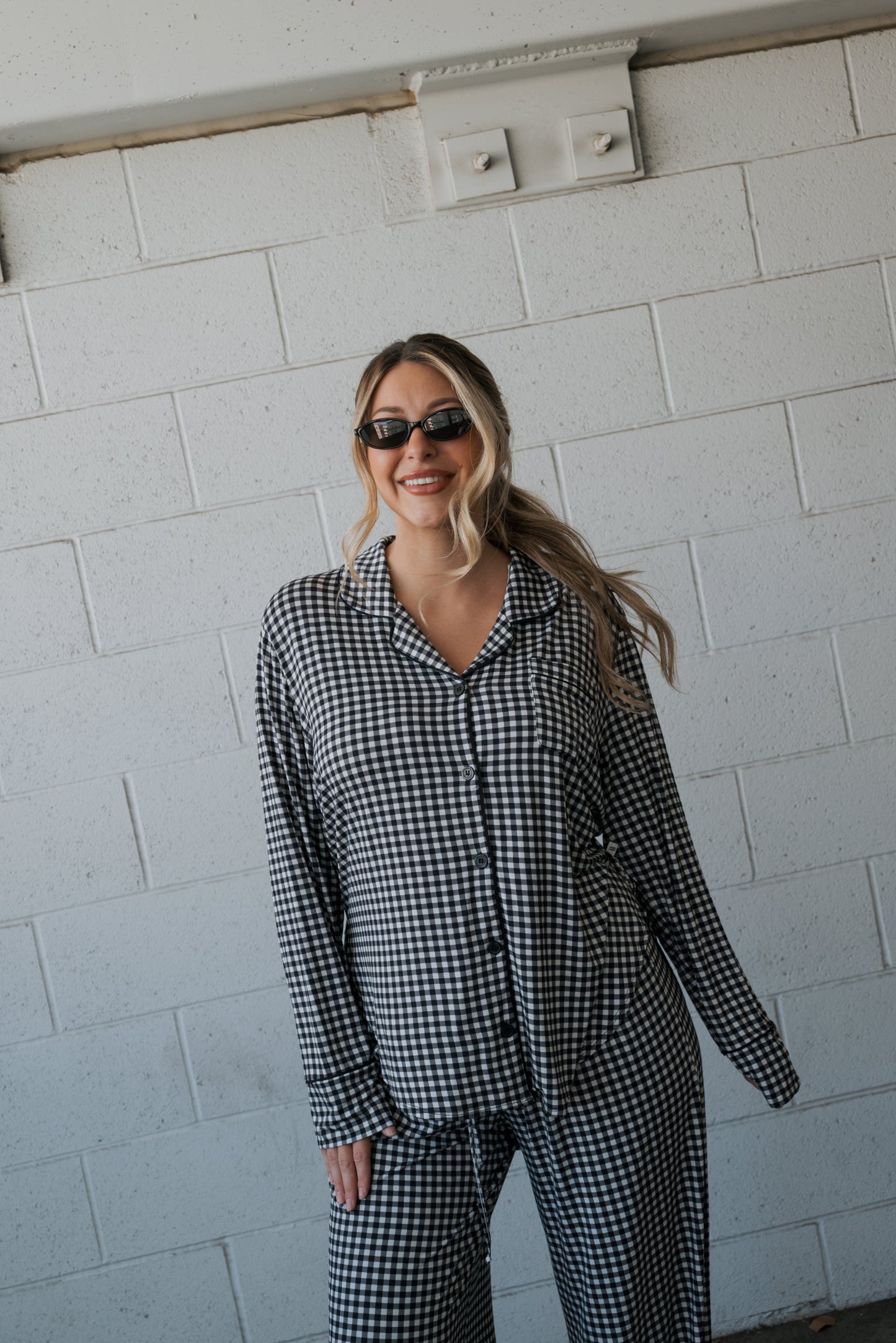 A woman wearing the forever french baby Women’s Bamboo Button Up Lounge Set in Black Gingham and sunglasses stands smiling in front of a white brick wall.