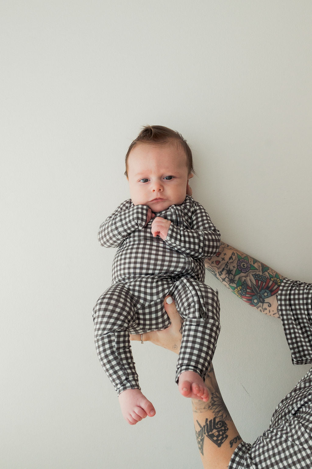 A baby in hypoallergenic Bamboo Zip Pajamas in Green Gingham by forever french baby is held up against a light background by an adult with colorful tattooed arms, whose shirt matches the baby’s outfit.
