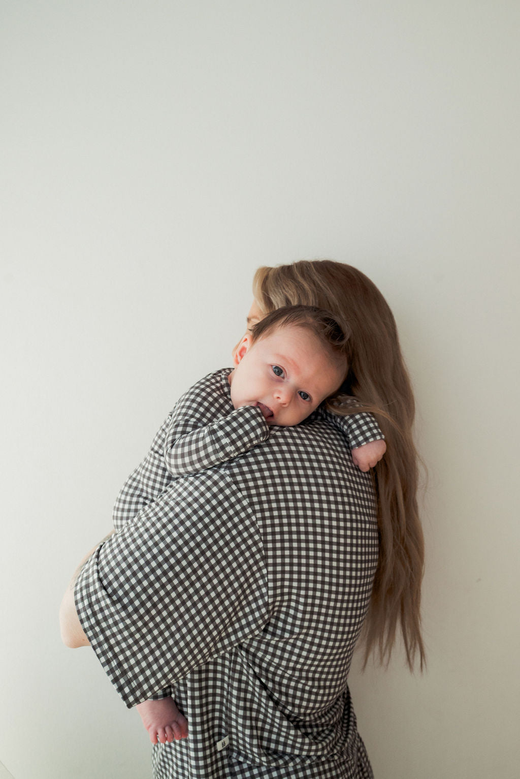 A baby in forever french baby's Bamboo Zip Pajamas in Green Gingham rests on the shoulder of an adult with long hair. Both wear matching checkered outfits against a white background, keeping the baby cozy while gazing at the camera.