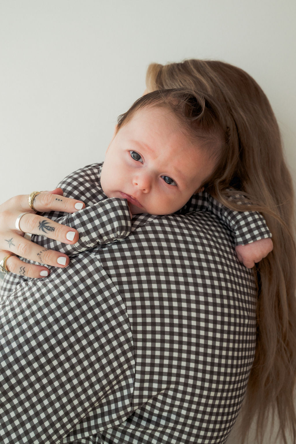 A baby in forever french baby's Bamboo Zip Pajamas | Green Gingham rests on an adult's shoulder, both dressed in matching gingham outfits as the adult’s tattooed, ring-adorned hand gently supports the baby.