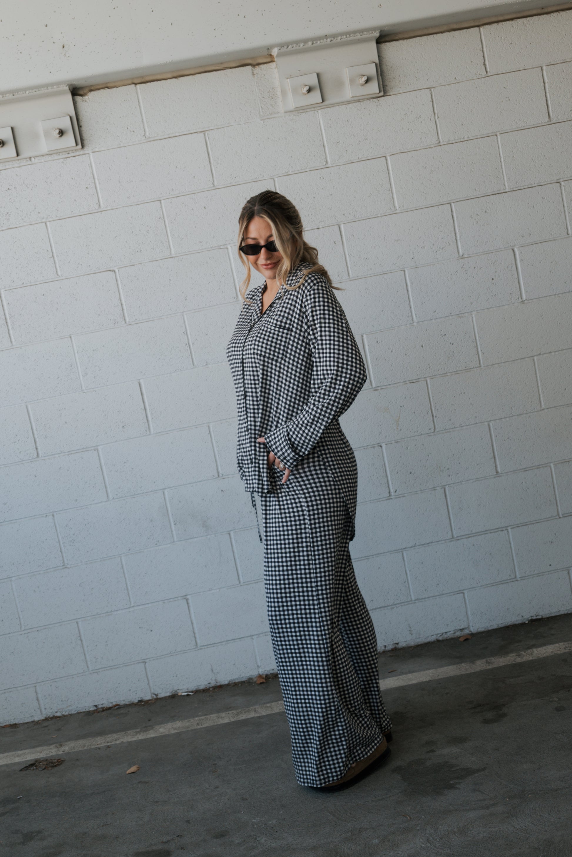 A person wearing sunglasses and the forever french baby Women’s Bamboo Button Up Lounge Set in Black Gingham stands sideways in front of a white brick wall, looking at the camera.
