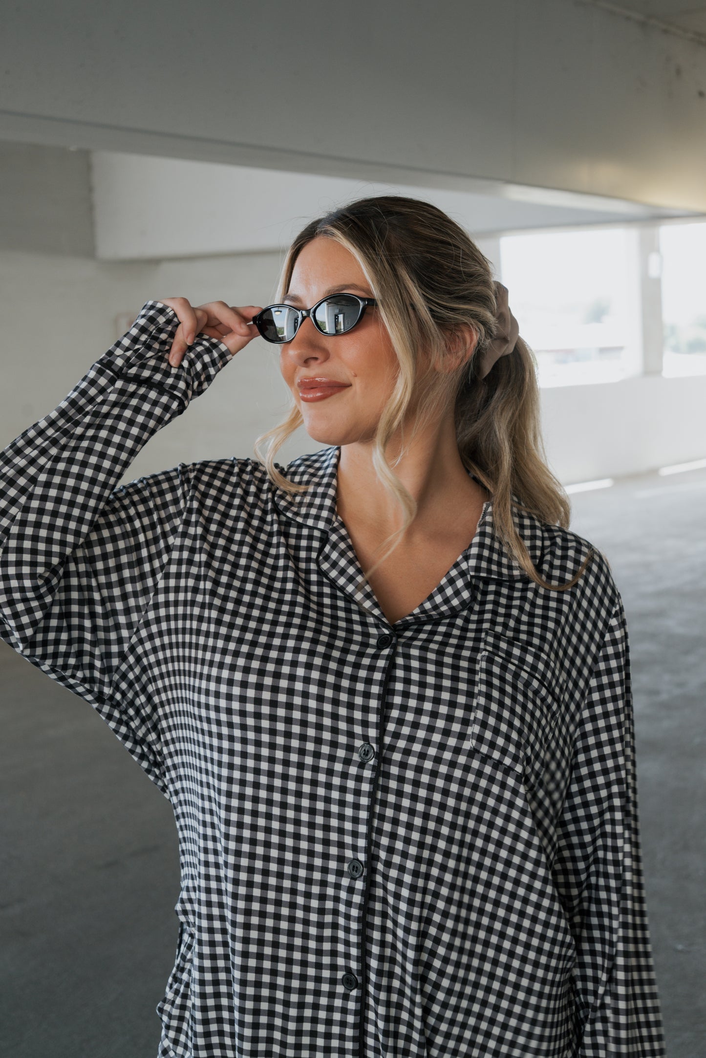 A woman in the forever french baby Women’s Bamboo Button Up Lounge Set in Black Gingham adjusts her sunglasses while standing in a bright, indoor parking area, effortlessly showcasing the comfort and relaxed style of bamboo clothing.