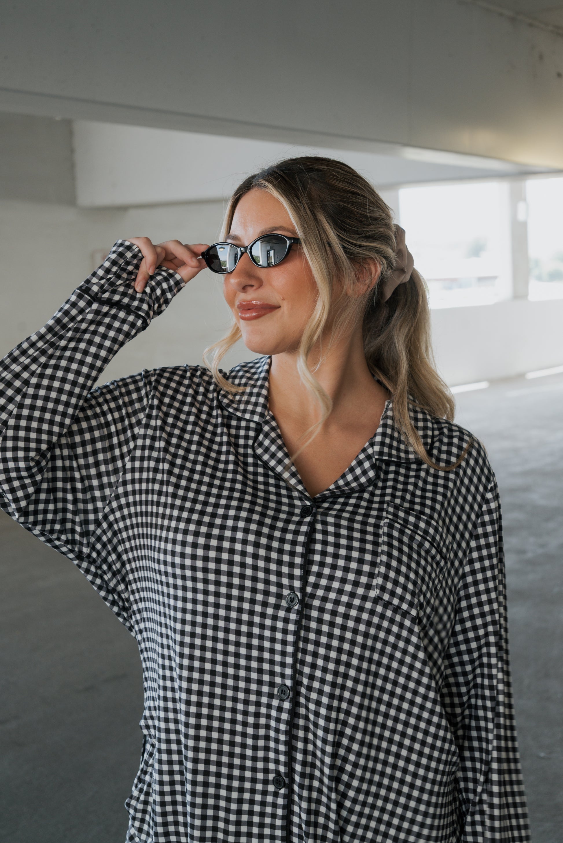 A woman in the forever french baby Women’s Bamboo Button Up Lounge Set in Black Gingham adjusts her sunglasses while standing in a bright, indoor parking area, effortlessly showcasing the comfort and relaxed style of bamboo clothing.
