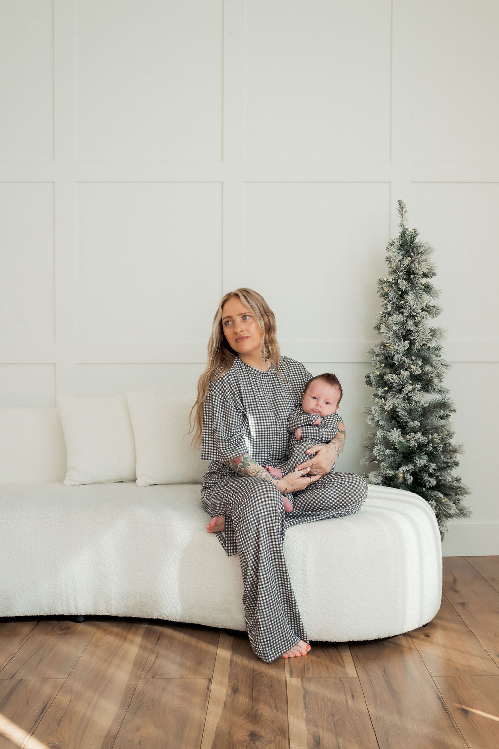 A woman in the forever french baby Women's Boxy Tee Lounge Set in Green Gingham sits barefoot on a white couch, holding a baby in matching hypo-allergenic pajamas. A small frosted Christmas tree is nearby in the bright, minimalist room.