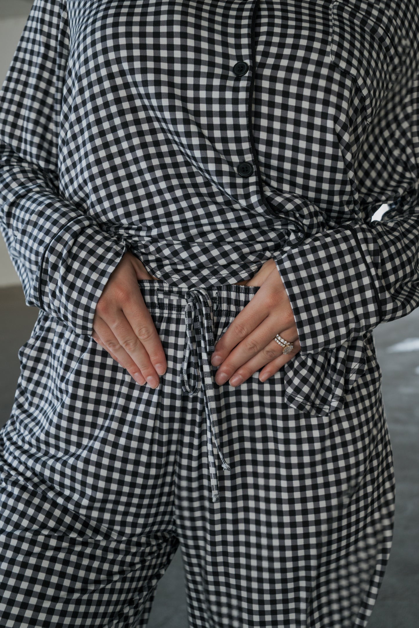 A person is shown wearing the Women’s Bamboo Button Up Lounge Set in Black Gingham by forever french baby, with hands resting on their stomach and rings adorning their left hand. The image focuses on the torso and lower part of the face, drawing attention to the soft bamboo material and relaxed fit of the wide-leg lounge pants.