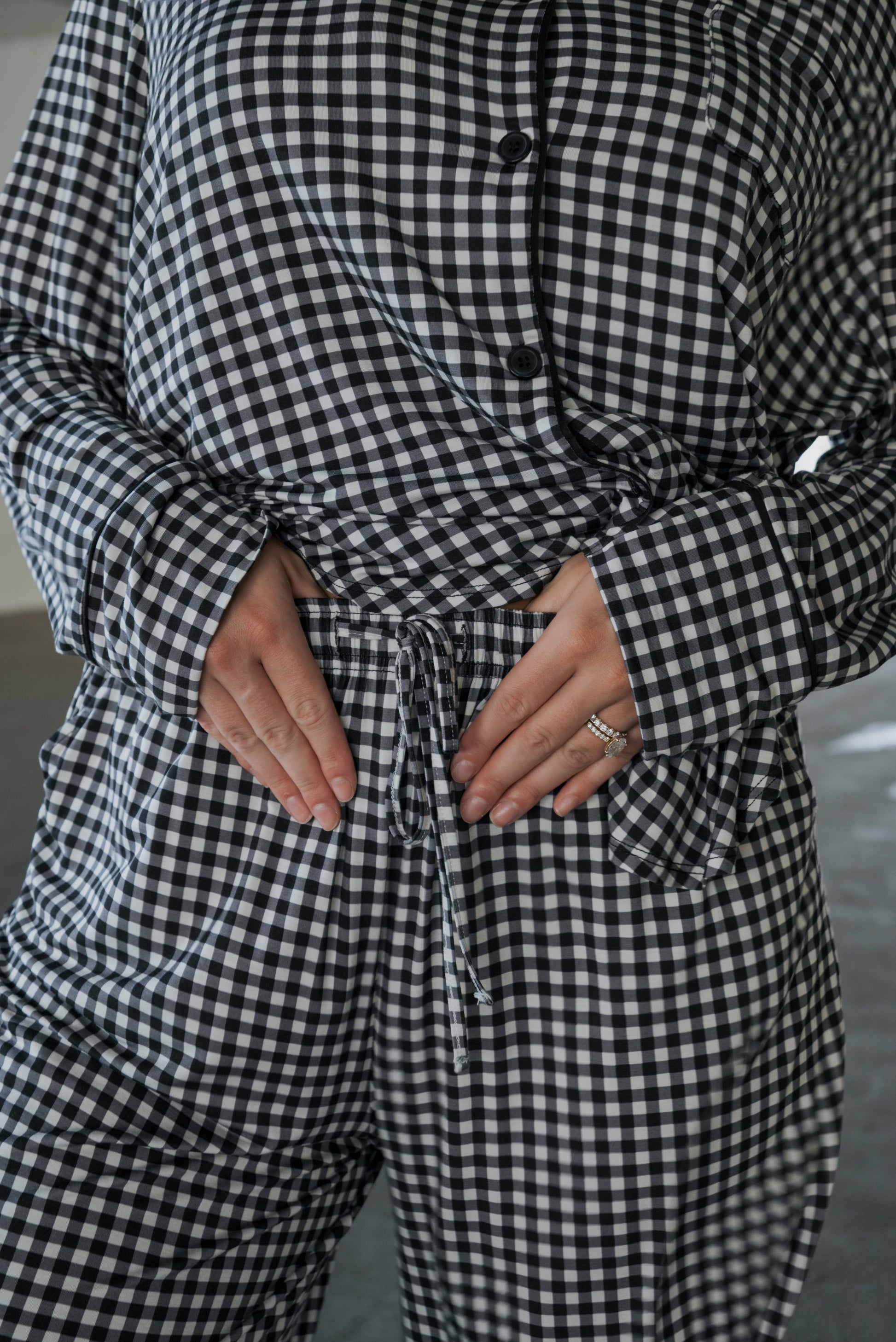 A person is shown wearing the Women’s Bamboo Button Up Lounge Set in Black Gingham by forever french baby, with hands resting on their stomach and rings adorning their left hand. The image focuses on the torso and lower part of the face, drawing attention to the soft bamboo material and relaxed fit of the wide-leg lounge pants.