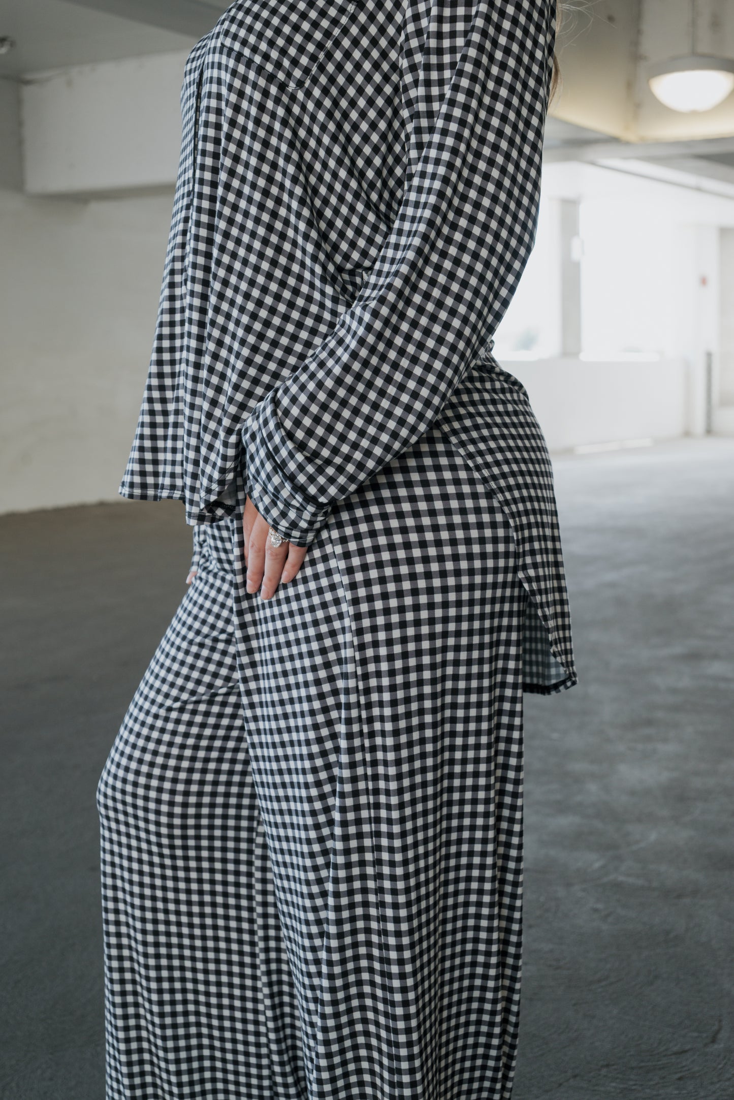A person wearing the forever french baby Women’s Bamboo Button Up Lounge Set in Black Gingham stands indoors with one hand on their hip. The background features an empty, white-walled space illuminated by natural light.