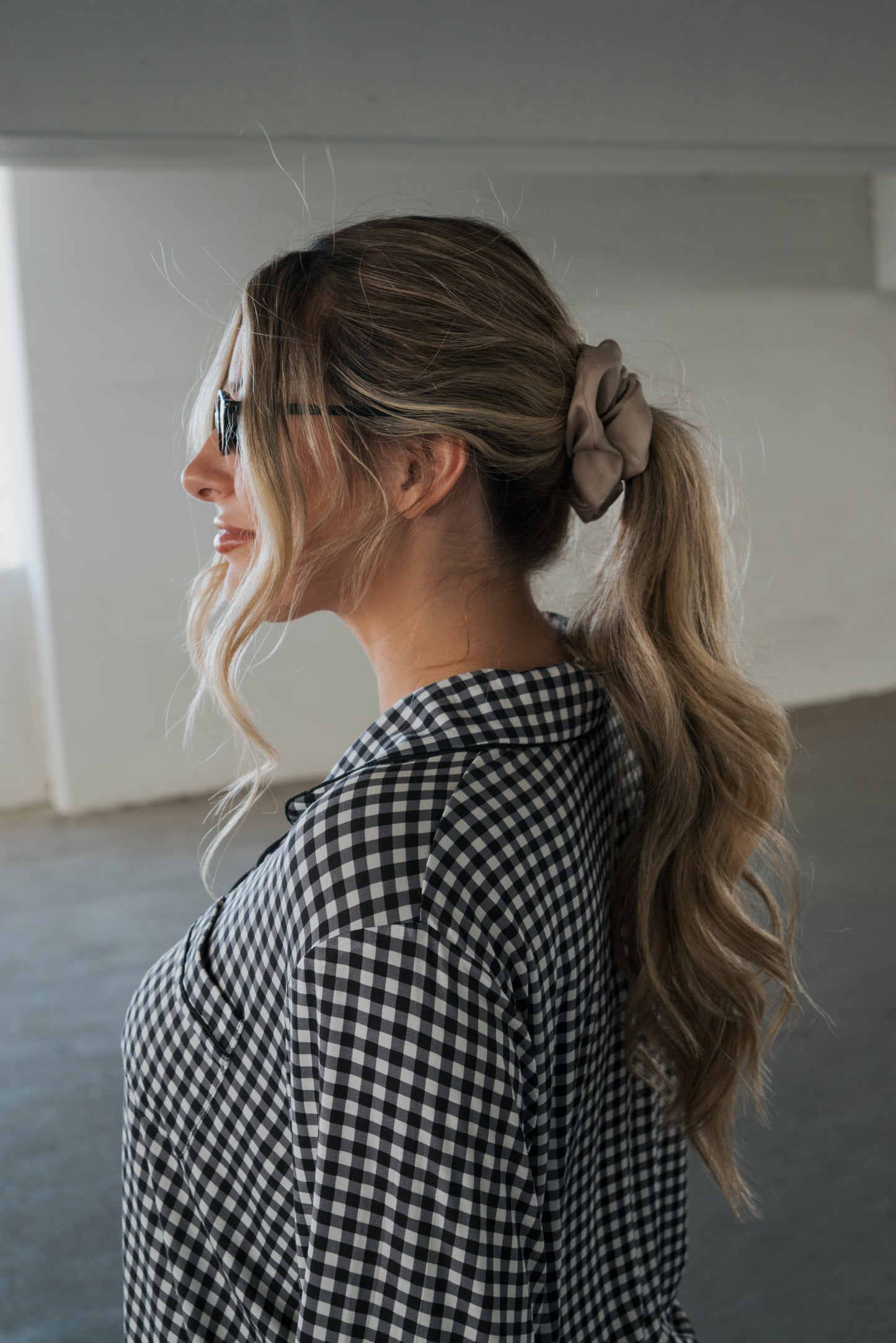 A woman with long wavy hair in a ponytail, secured by a beige scrunchie and wearing sunglasses, stands indoors facing left. She is dressed in the forever french baby Women’s Bamboo Button Up Lounge Set in Black Gingham.