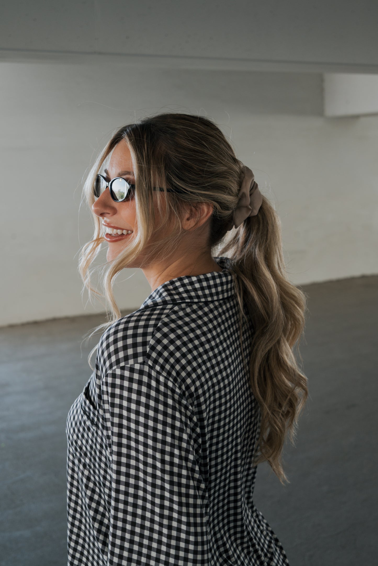 A woman with long wavy blonde hair in a ponytail, wearing sunglasses and the forever french baby Women’s Bamboo Button Up Lounge Set in Black Gingham, smiles and looks over her shoulder in a bright indoor space, effortlessly embodying the relaxed elegance of soft bamboo clothing.