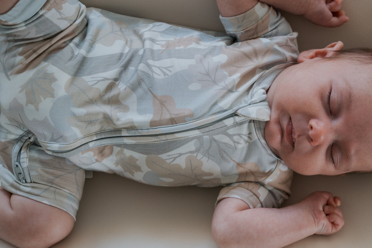 A baby sleeps peacefully on their back, wearing the Shortie Bamboo Zip | Woodland romper by forever french baby—a light-colored, leaf-patterned zip-up crafted from hypoallergenic bamboo sleepwear material.