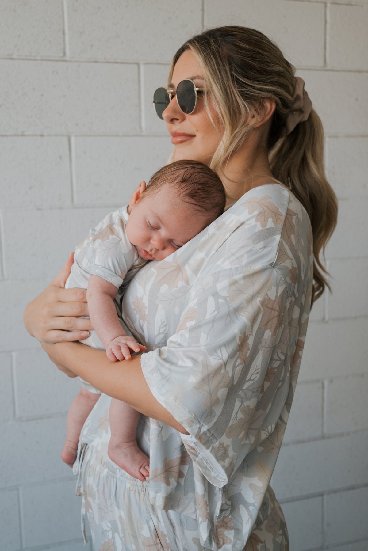 Wearing the Women’s Bamboo Crop Tee Lounge Set in Woodland from forever french baby, a woman in sunglasses holds a sleeping baby against her chest. Both are dressed in light-colored, floral-patterned clothes made from soft bamboo fabric and stand before a white brick wall.