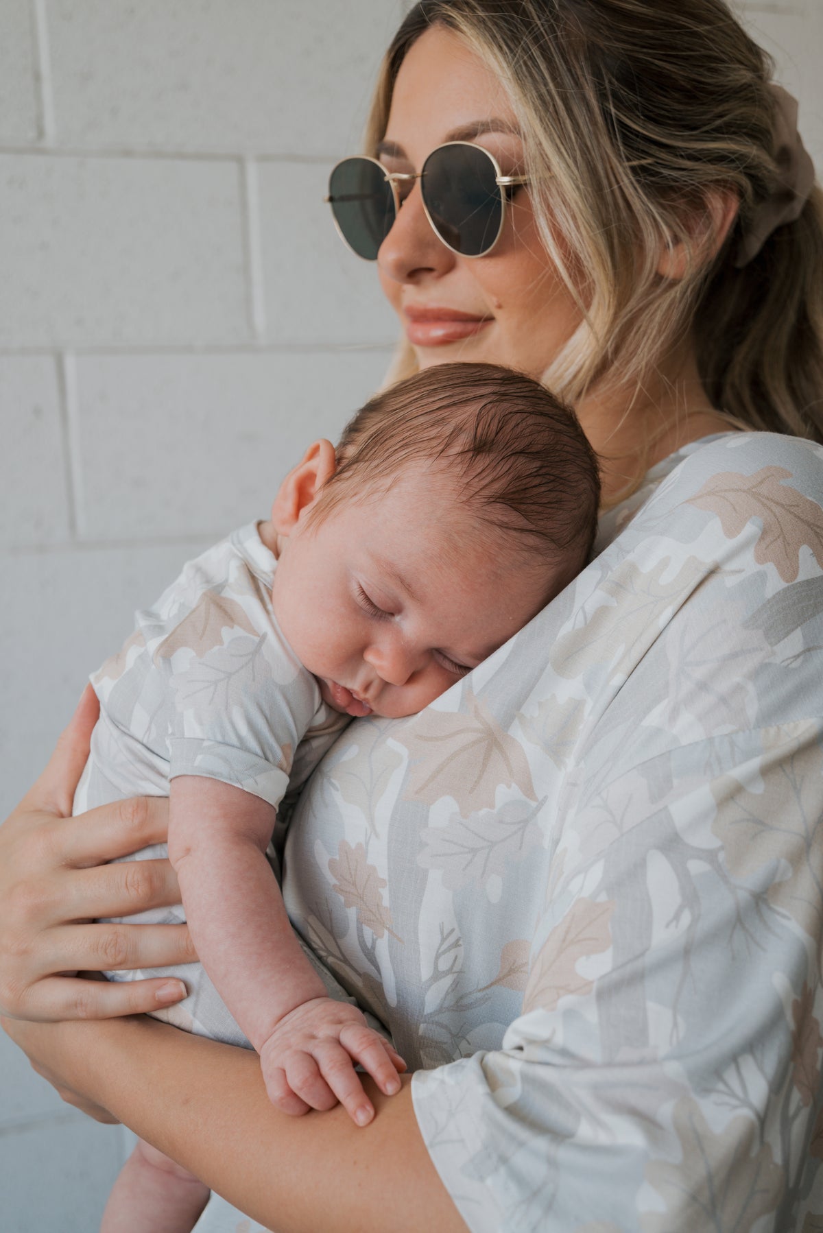A woman wearing sunglasses smiles gently while holding a sleeping baby against her chest. Both are dressed in matching forever french baby outfits, featuring the Shortie Bamboo Zip | Woodland—a cozy, hypoallergenic romper with a nature-inspired pattern that’s perfect for sensitive skin.