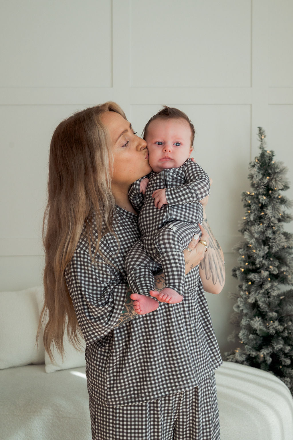 A woman wears the forever french baby Women's Boxy Tee Lounge Set in Green Gingham and kisses a baby in matching pajamas, holding them upright with a small decorated Christmas tree in the background.